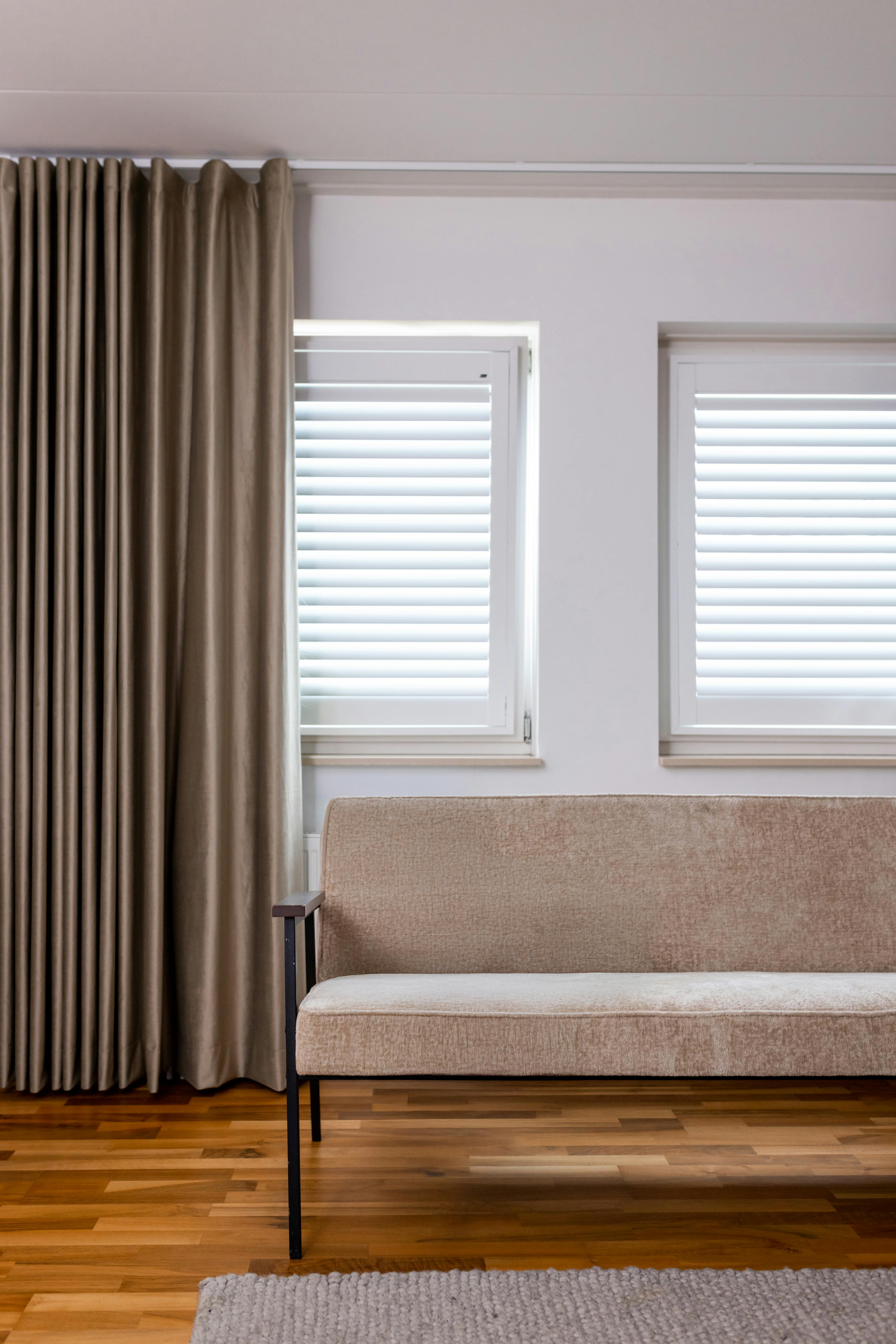 Witte shutters op ramen in een lichte woonkamer met een beige fauteuil