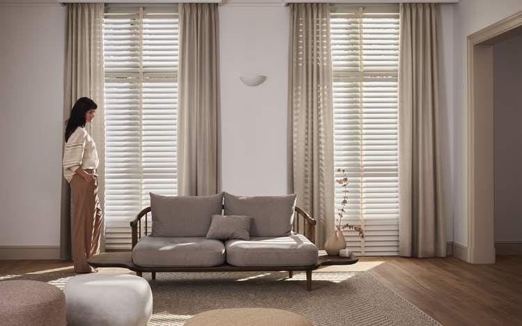 Een vrouw die voor ramen staat met beige gordijnen en witte jaloezieën in een gezellige woonkamer