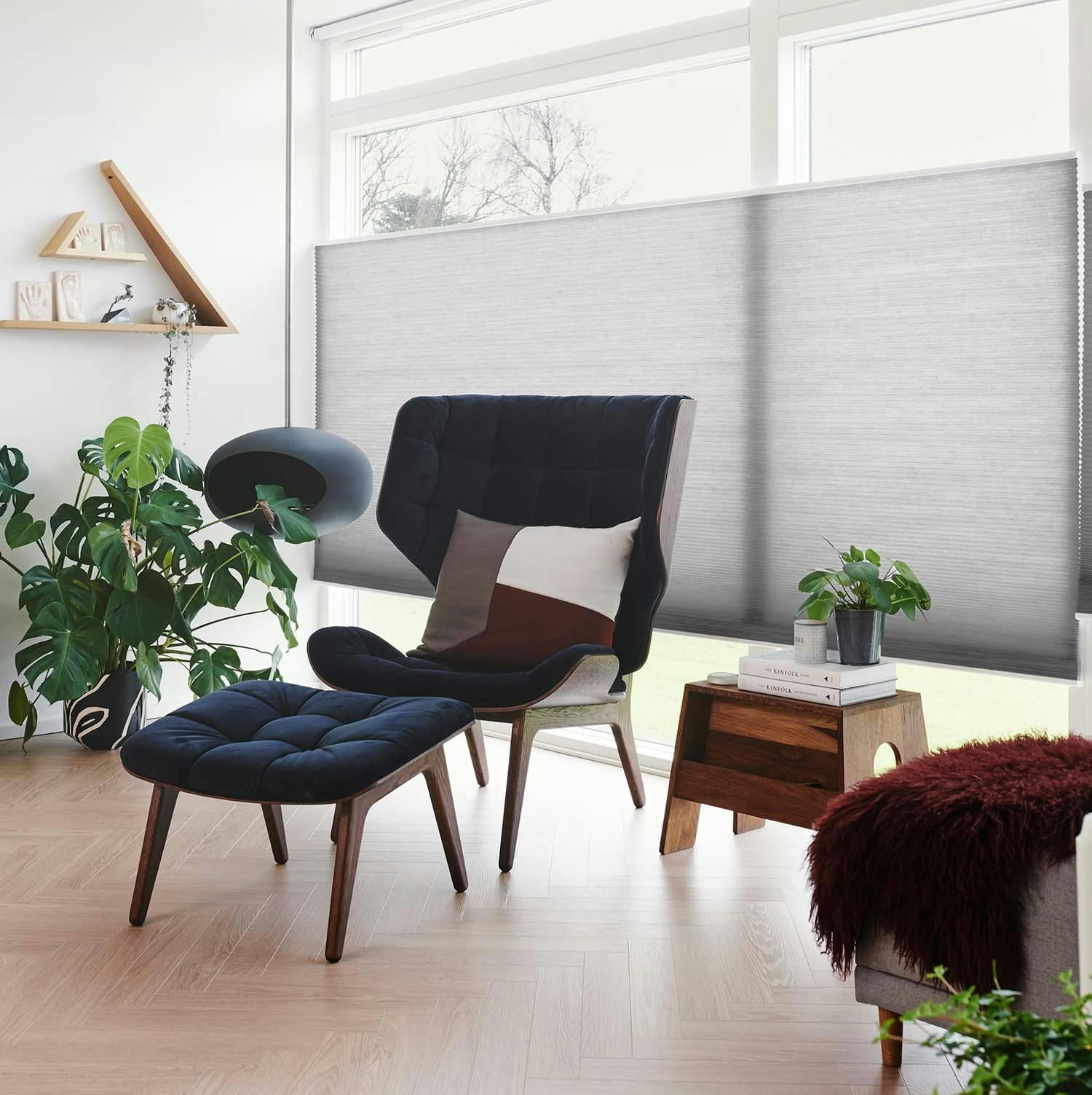 Modernt vardagsrum med Duette®-gardin i grått, svart fåtölj med fotpall, färgglad kudde, grön växt, träbord med böcker och krukväxt, samt dekorativa detaljer på hyllan.