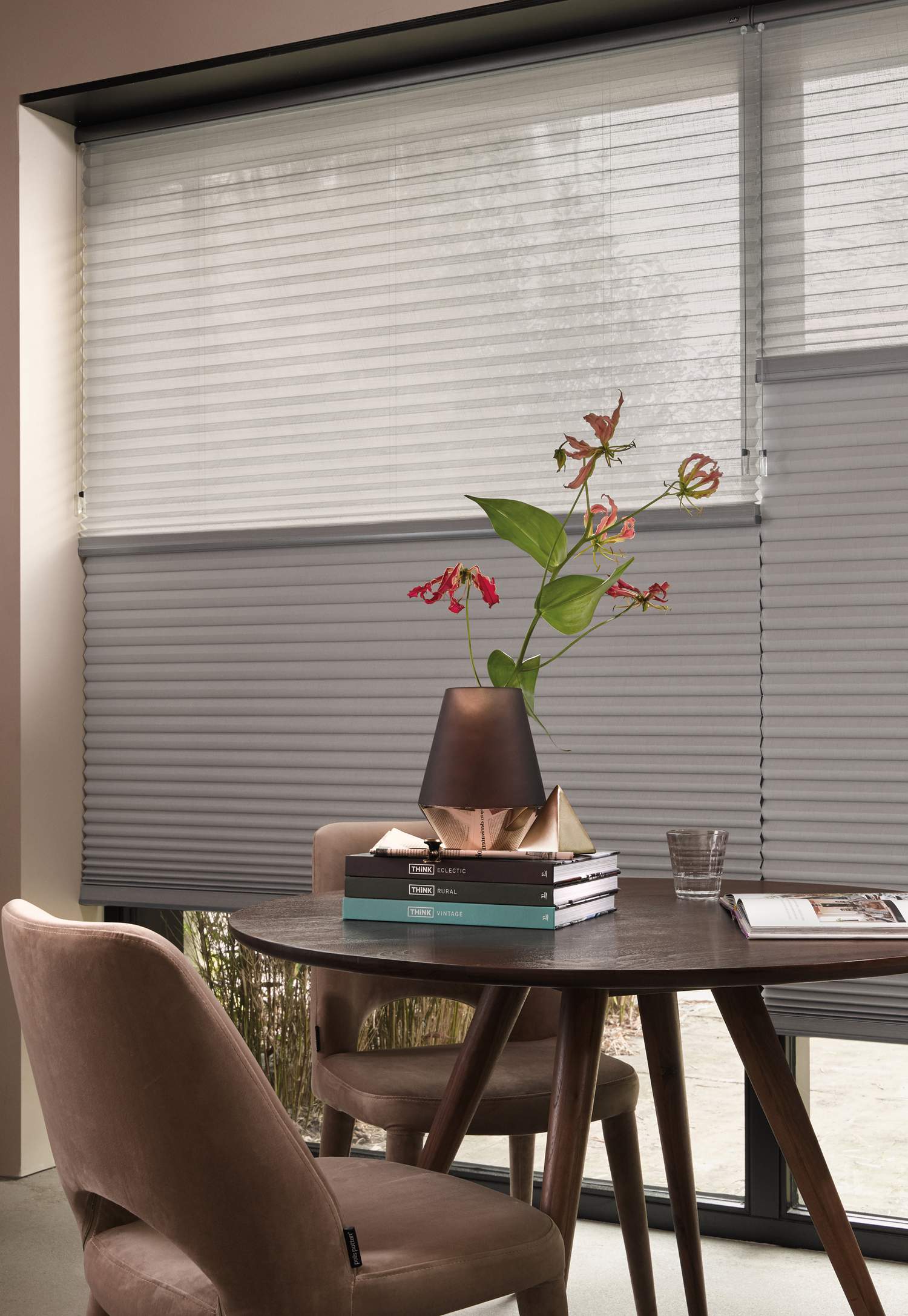 Rundt bord med vase, bøker og glass foran store vinduer med persienner.