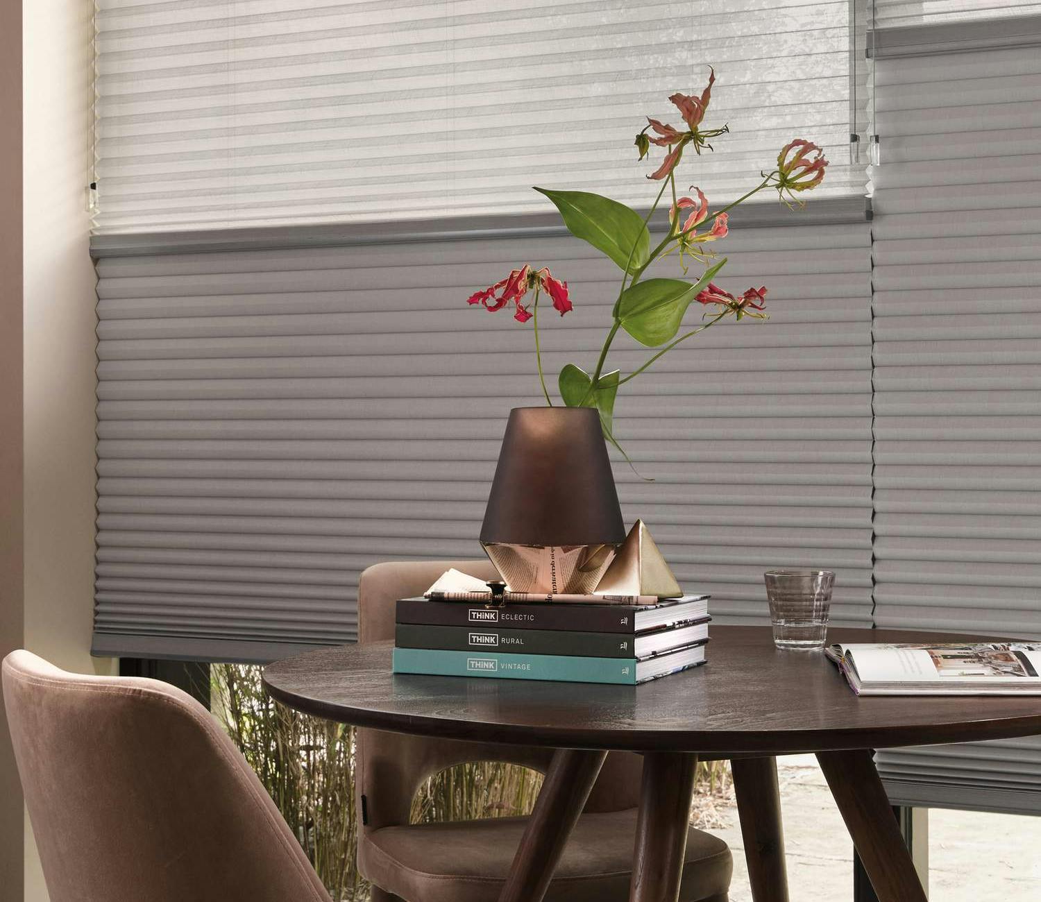 Rundt bord med vase, bøker og glass foran store vinduer med persienner.