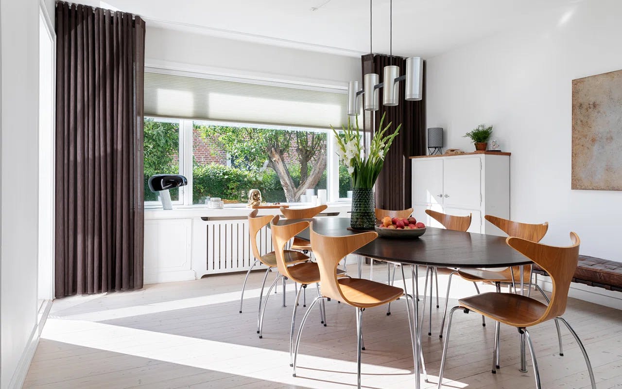 Spisestue med mørkt trebord, vindu med persienner og utsikt mot hage.