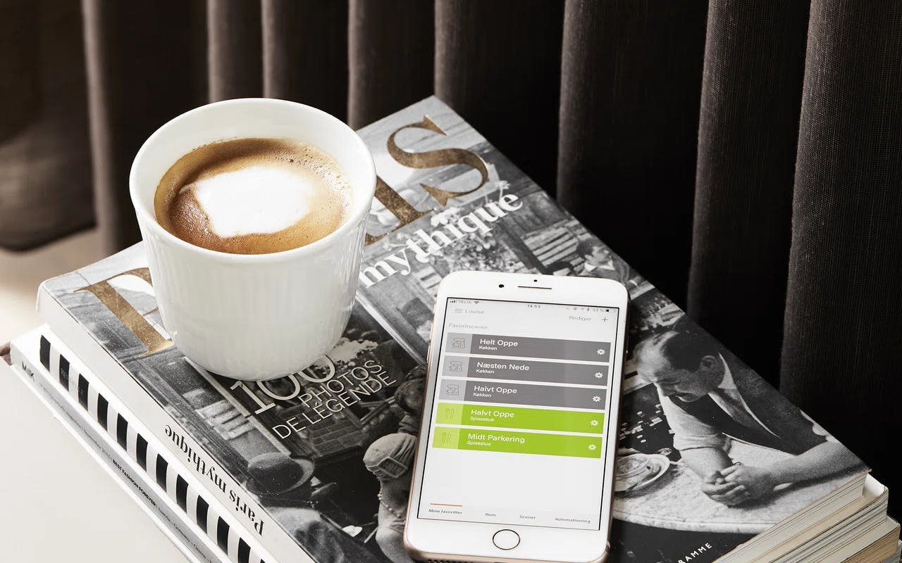 Kaffekopp med skum på toppen plassert på en bok med tittelen 'PARIS', ved siden av en smarttelefon som viser en app med hotellvalg.