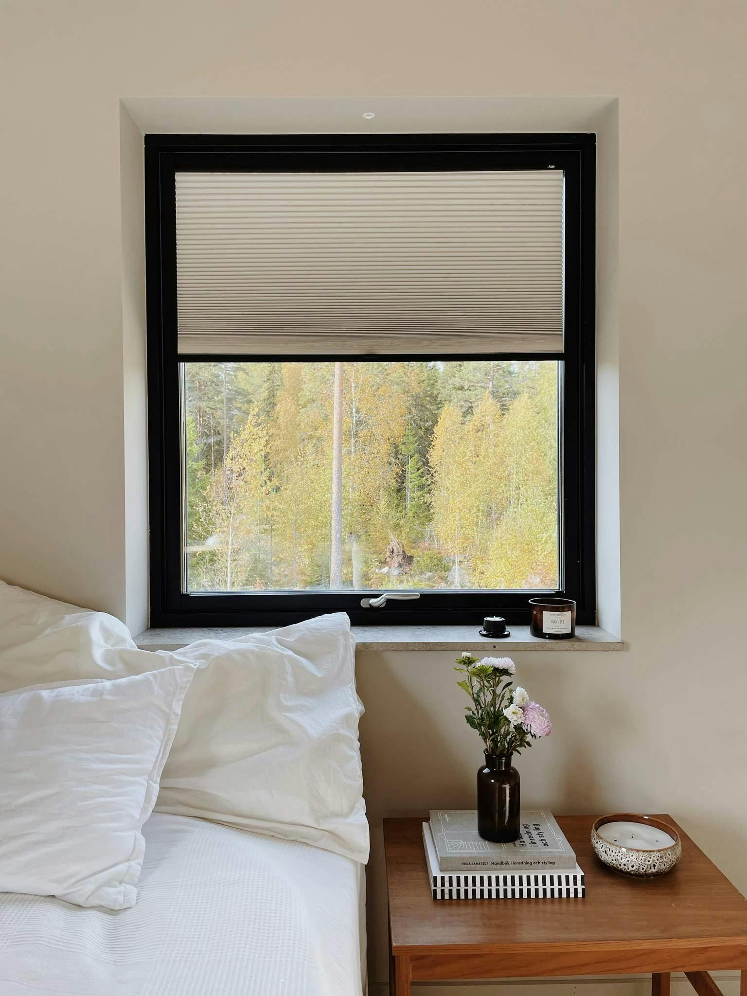 Soverom med vindu og utsikt mot høsttrær, seng med hvitt sengetøy og nattbord med bøker, vase og dekor.