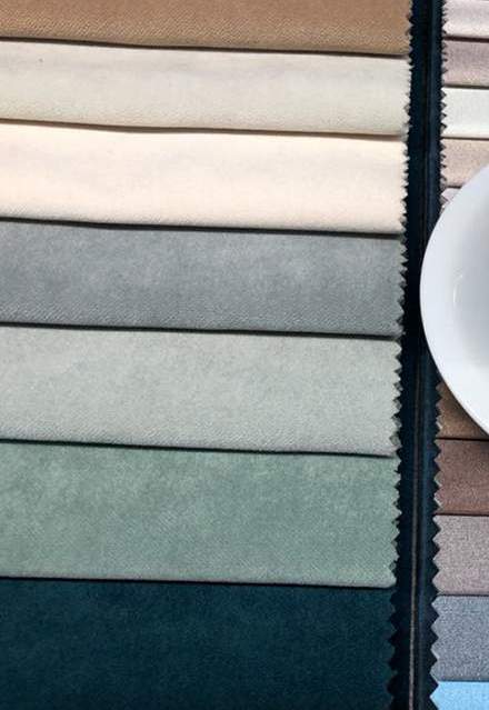 Ett urval av tygprover ordnade i en färgskala från ljusa till mörka nyanser, från beige och kräm till grått, grönt och mörk teal. Proverna har sicksackkanter och ligger prydligt. Till höger syns en del av en vit tallrik.