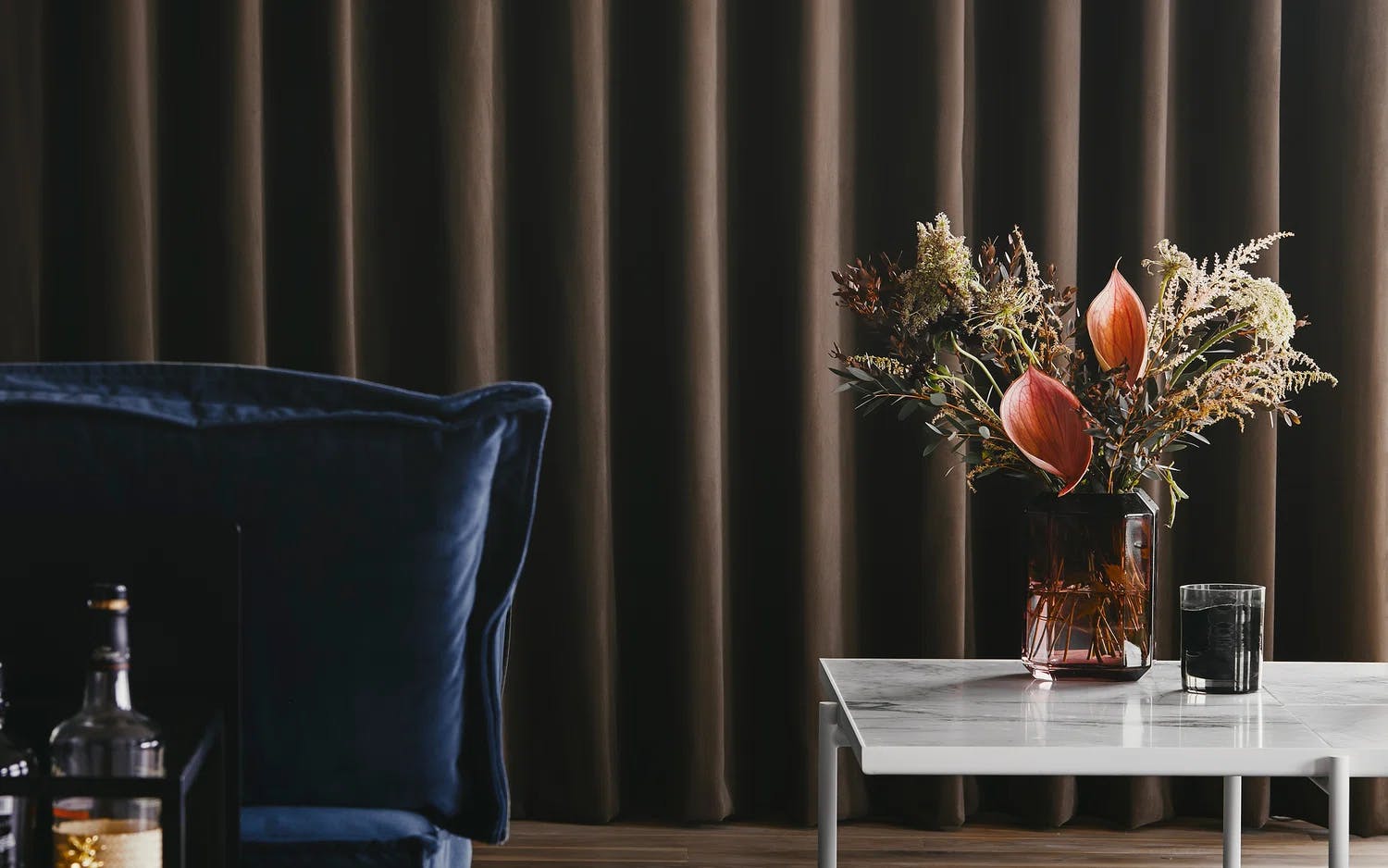 Ett rum med mörka gardiner i bakgrunden. Till vänster finns en blå fåtölj och en hylla med flaskor. Till höger står ett vitt marmorbord med en vas fylld med torkade blommor och växter, samt ett litet svart ljus.