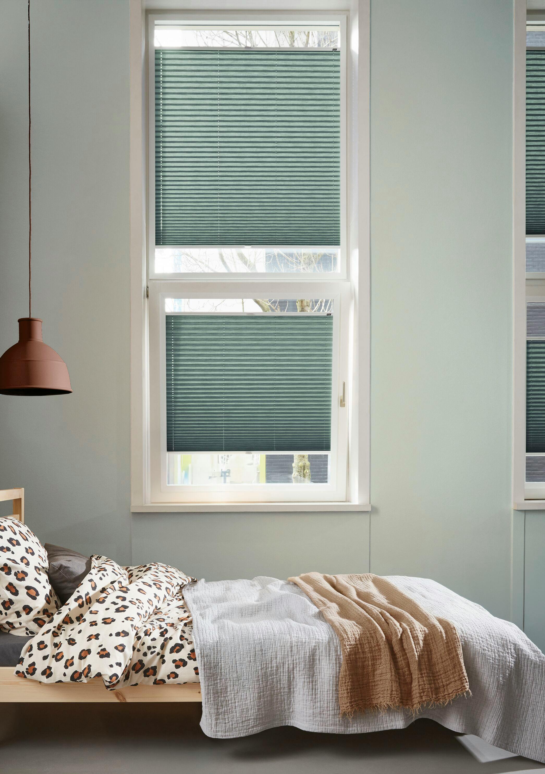 Two green plisse blinds on tilt and turn windows next to a stylish bed