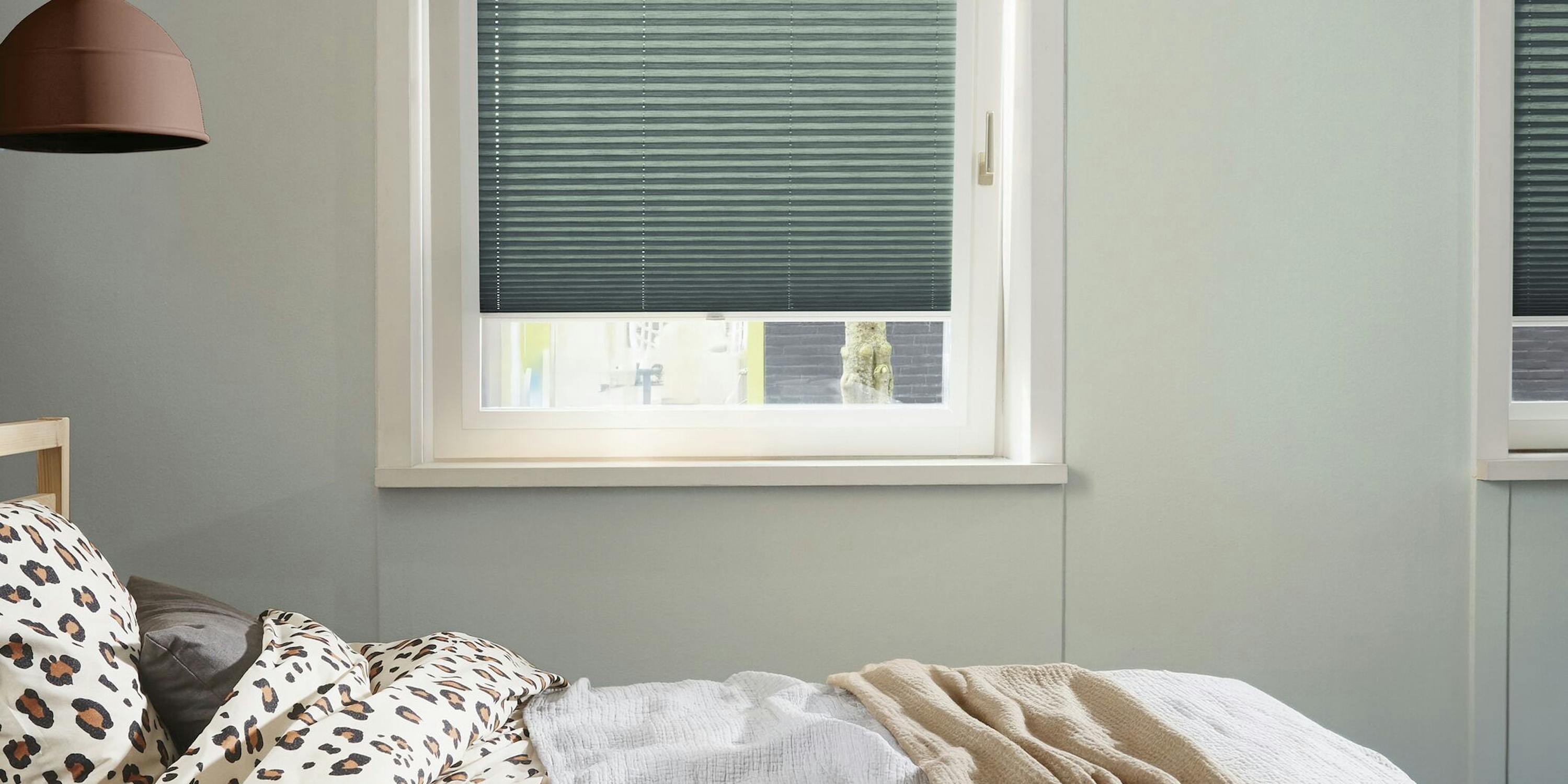 Two green plisse blinds on tilt and turn windows next to a stylish bed