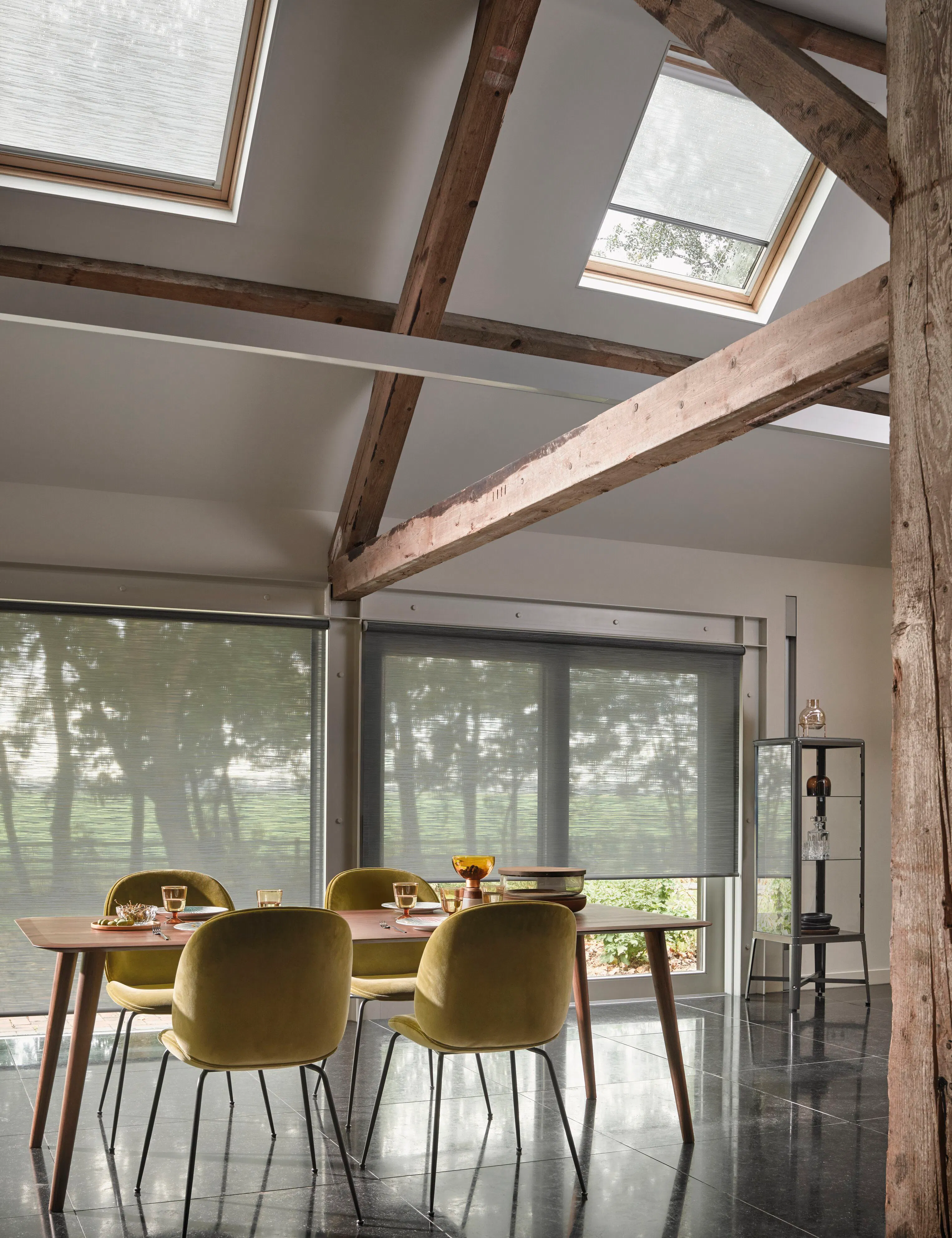 Two grey roller blinds on big bifold doors in a dining room under a slanted roof with two windows featuring day and night skylight blinds