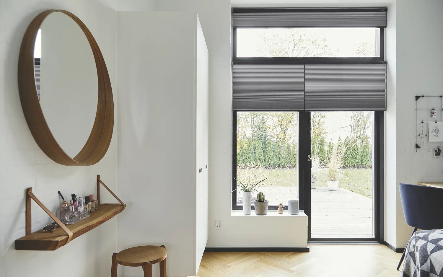 Ett modernt, minimalistiskt rum med ett stort fönster och dörr som leder ut till en altan med krukväxter och grönska. På väggen sitter en rund spegel med träram ovanför en trähylla med småsaker, samt en liten träpall under. Till höger finns ett högt vitt skåp och en del av ett skrivbord med väggdekorationer som foton och anteckningar på ett metallnät.