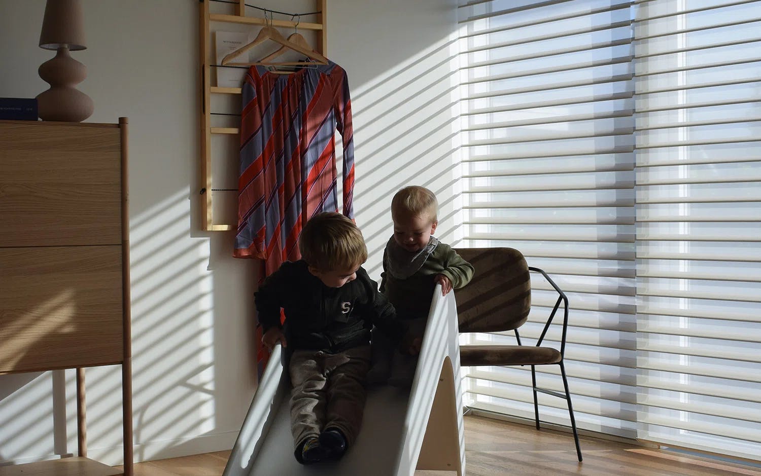 Ett modernt hemmakontor eller arbetsrum med stora fönster täckta av mörka rullgardiner. Ett träbord står i mitten med en laptop, böcker, en flaska och glas. Runt bordet finns stolar och längs väggen en sideboard med fler böcker och krukväxter. Utanför fönstren syns en grönskande trädgård.