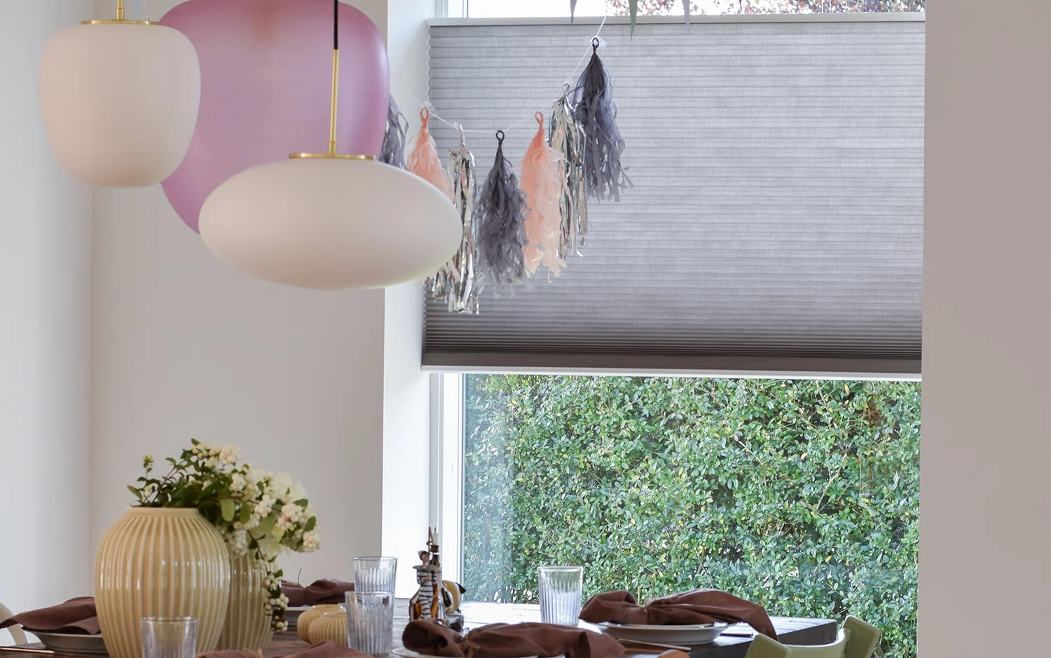 En matplats med ett dukat bord med tallrikar, glas, servetter och en vas med blommor. Två taklampor hänger ovanför bordet. I bakgrunden finns ett fönster med en grå rullgardin och dekorativa tofsar, samt grönska utanför.