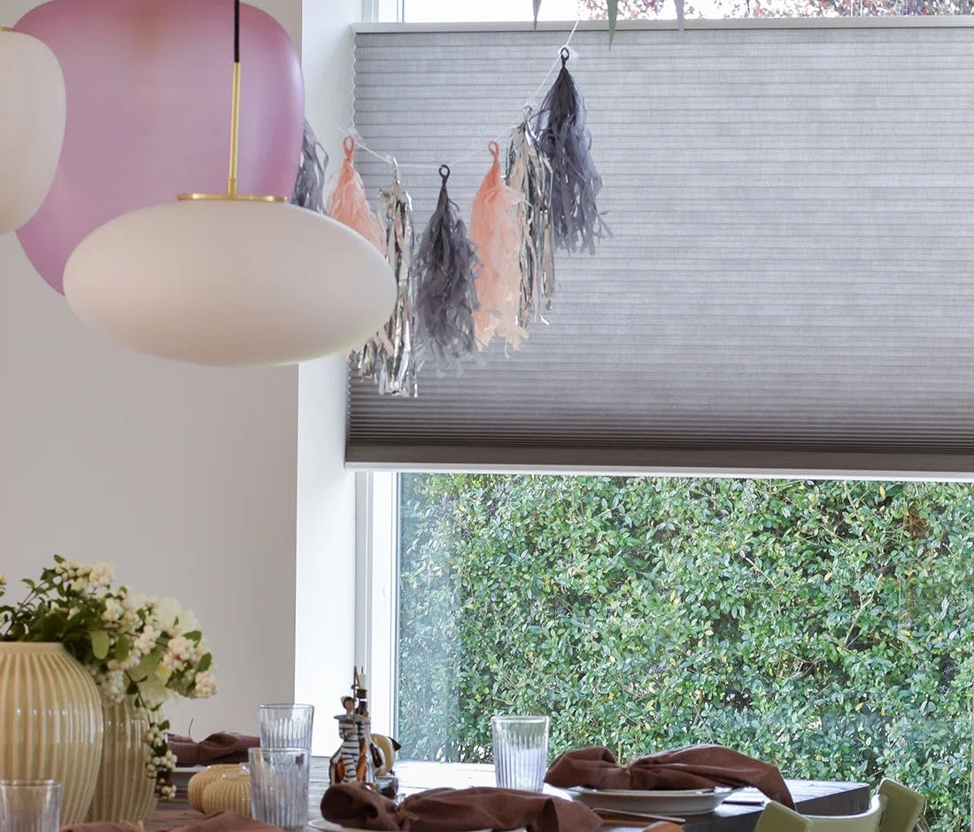 En matplats med ett dukat bord med tallrikar, glas, servetter och en vas med blommor. Två taklampor hänger ovanför bordet. I bakgrunden finns ett fönster med en grå rullgardin och dekorativa tofsar, samt grönska utanför.