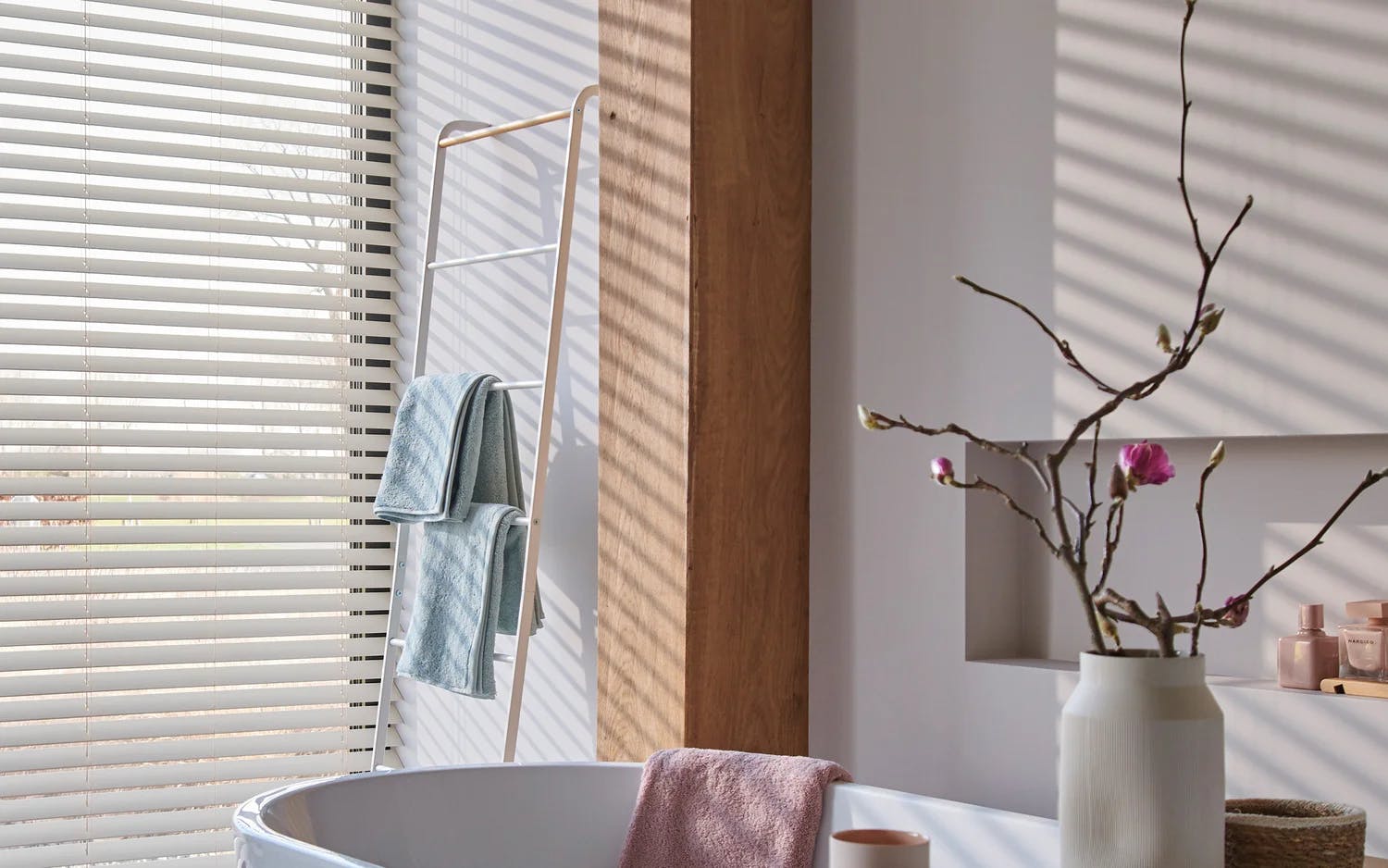 Ett lugnt badrum med ett vitt badkar och en rosa handduk hängande över kanten. Till vänster står ett fönster med horisontella persienner som släpper in solljus och skapar skuggmönster. En trästege fungerar som handdukshängare och håller två vikta handdukar i blått och grönt. Till höger står en vit vas med rosa blommande kvistar på en hylla. Rummet har ljusa väggar och trädetaljer som ger en minimalistisk och rofylld känsla.
