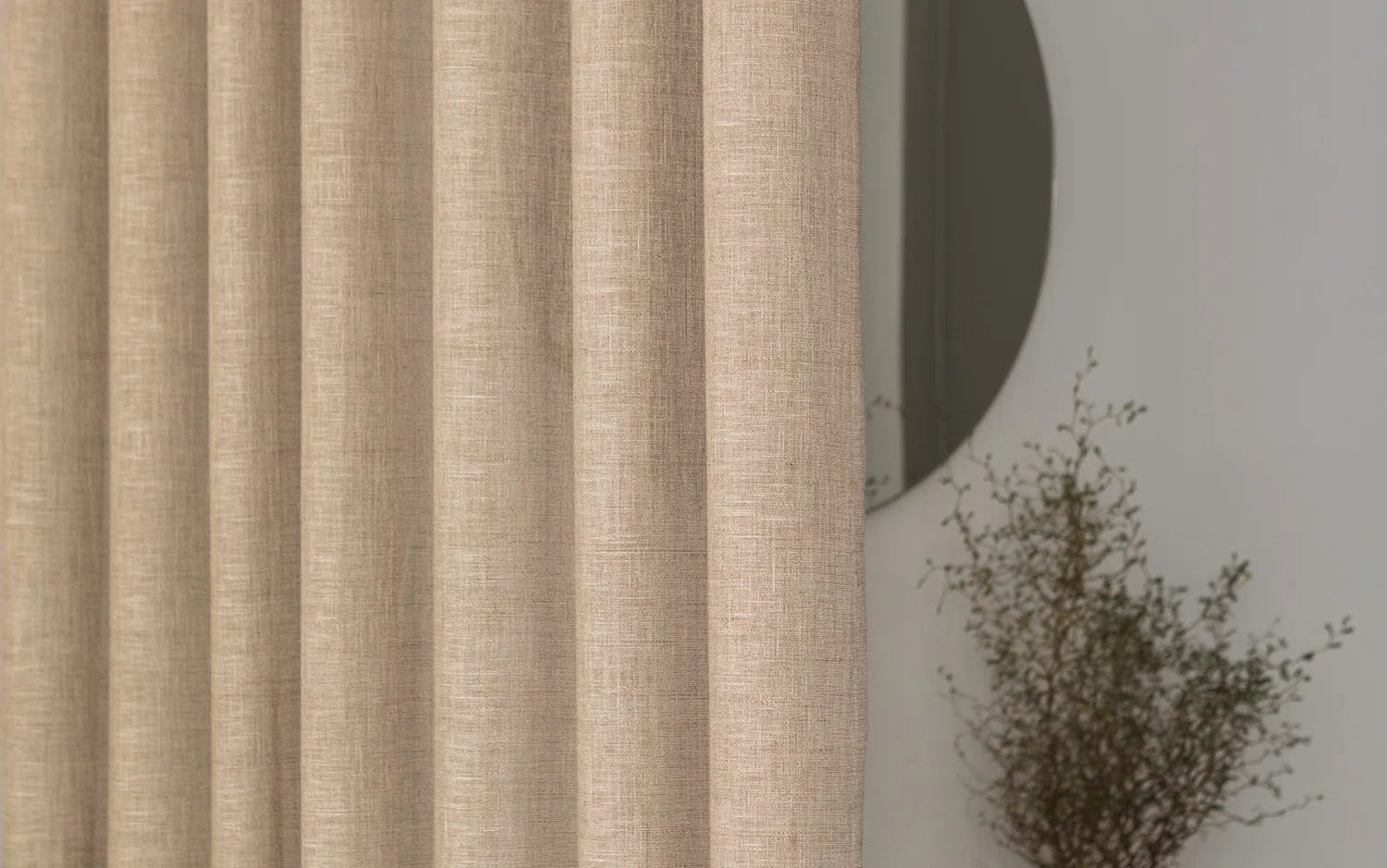 Närbild på beige gardiner med texturerat tyg. I bakgrunden syns en rund spegel på väggen och en växt med tunna grenar och små blad.