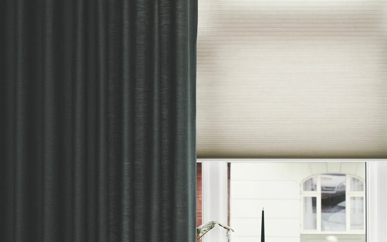 Delvis vy av ett fönster med två olika typer av fönsterbehandlingar: till vänster en mörk gardin med vertikala veck och till höger en ljus plisségardin. Fönstret har vit karm och utanför syns en byggnad med krämfärgade väggar och välvda fönster.