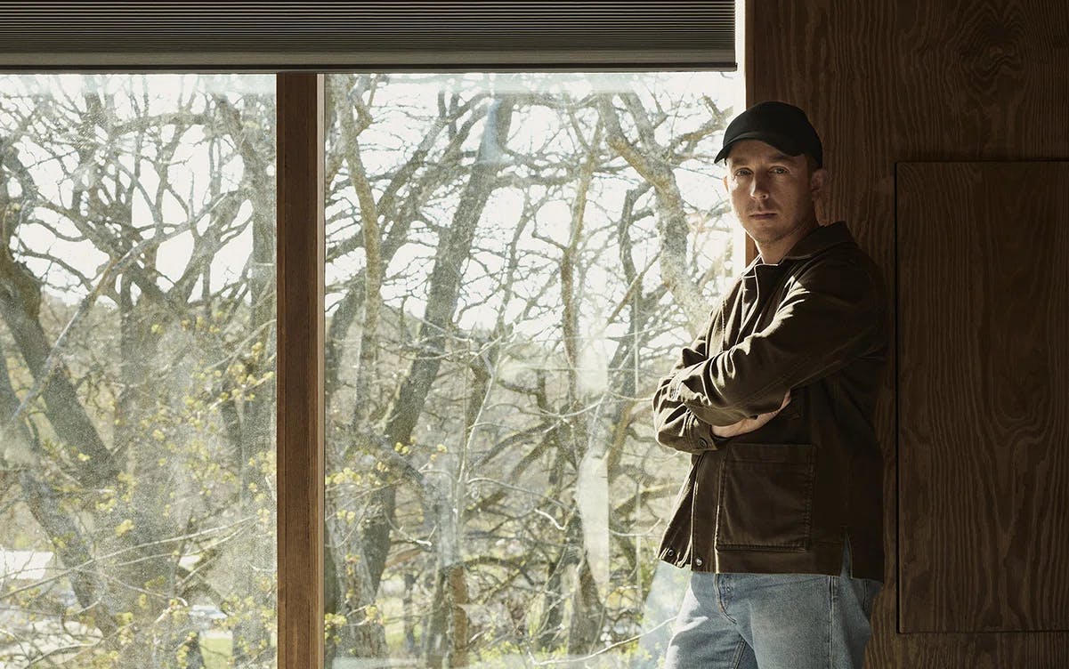 En person står inomhus intill ett stort fönster, iförd mörk jacka, ljusblå jeans och mörk keps. Personen lutar sig mot en trävägg med armarna i kors. Utanför fönstret syns ett naturlandskap med träd som börjar knoppas, vilket tyder på tidig vår. Bilden fångar kontrasten mellan den varma inomhusmiljön och det svala, naturliga utomhuslandskapet.