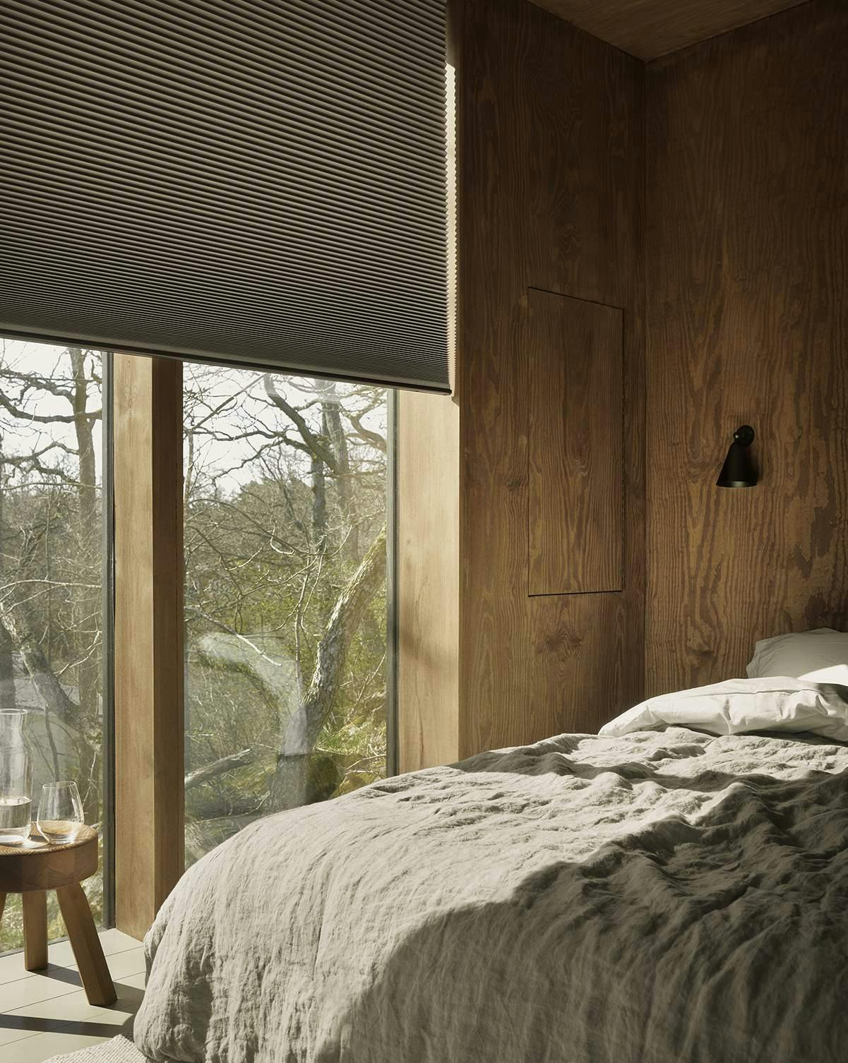 Ett mysigt sovrum med träväggar och stora fönster. Sängen har ett skrynkligt, ljusfärgat täcke och kuddar. Bredvid sängen står ett runt sidobord i trä med ett vattenglas och en karaff. Fönstret har delvis neddragna persienner som släpper in naturligt ljus och ger utsikt mot träd utanför. Miljön känns varm och inbjudande med en harmonisk koppling mellan inne och ute.