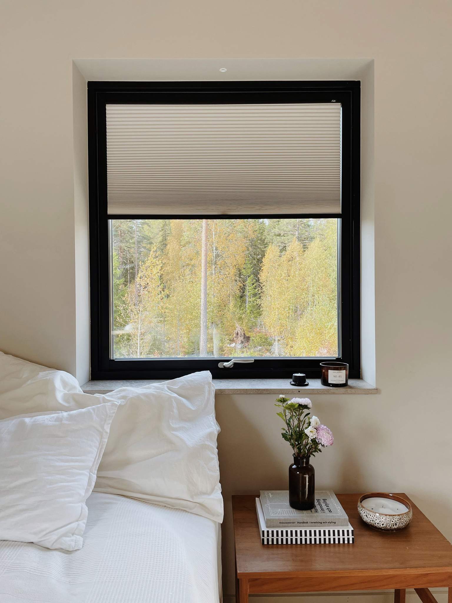 Ett mysigt sovrum med ett fönster som har svart ram och delvis öppna persienner. Utanför syns höstträd med gula löv. Inne i rummet står en säng med vita kuddar och sängkläder till vänster. Bredvid sängen finns ett nattduksbord i trä med två staplade böcker, en liten vas med blommor och en dekorativ skål. Rummet har en lugn och inbjudande atmosfär med naturligt ljus och neutrala färger.