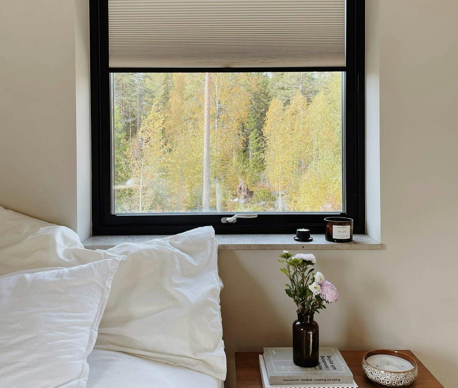 Ett mysigt sovrum med ett fönster som har svart ram och delvis öppna persienner. Utanför syns höstträd med gula löv. Inne i rummet står en säng med vita kuddar och sängkläder till vänster. Bredvid sängen finns ett nattduksbord i trä med två staplade böcker, en liten vas med blommor och en dekorativ skål. Rummet har en lugn och inbjudande atmosfär med naturligt ljus och neutrala färger.