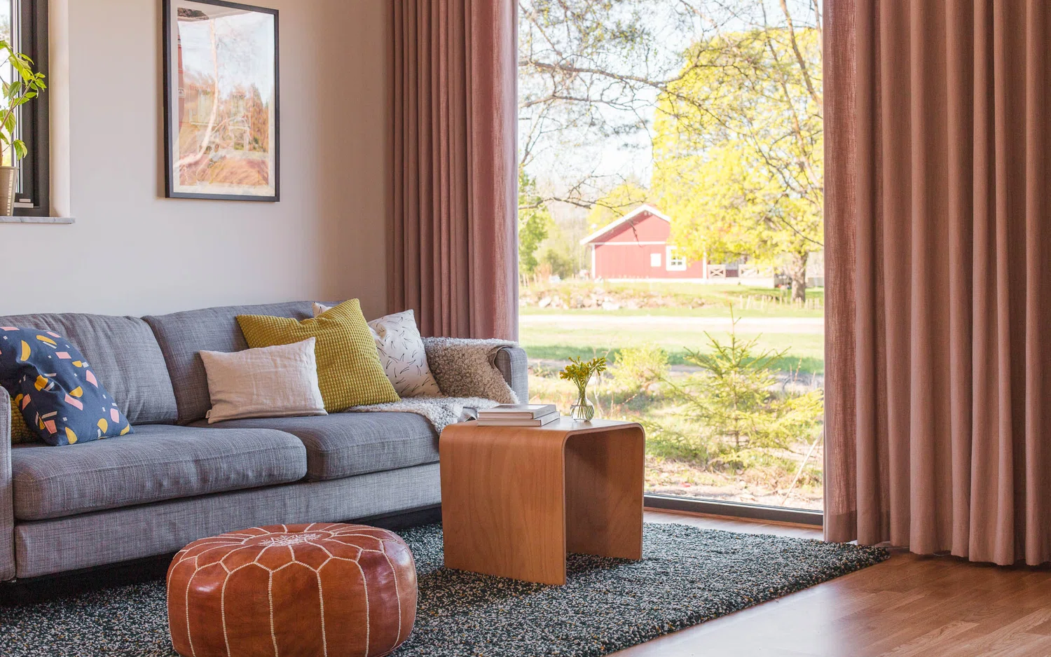Ett mysigt vardagsrum med en grå soffa fylld med kuddar i olika färger och mönster. Framför soffan står ett soffbord i trä med böcker och en liten vas med blommor. På golvet ligger en texturerad matta och en brun läderpuff. Ett stort fönster med rosa gardiner drar blicken mot utsikten utanför – gräs, träd och en röd byggnad i fjärran.