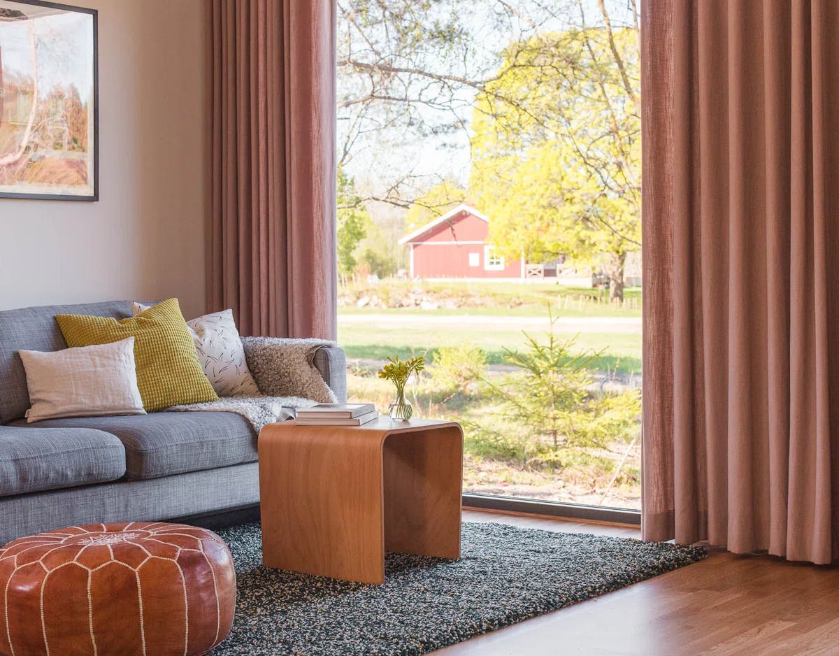 Ett mysigt vardagsrum med en grå soffa fylld med kuddar i olika färger och mönster. Framför soffan står ett soffbord i trä med böcker och en liten vas med blommor. På golvet ligger en texturerad matta och en brun läderpuff. Ett stort fönster med rosa gardiner drar blicken mot utsikten utanför – gräs, träd och en röd byggnad i fjärran.