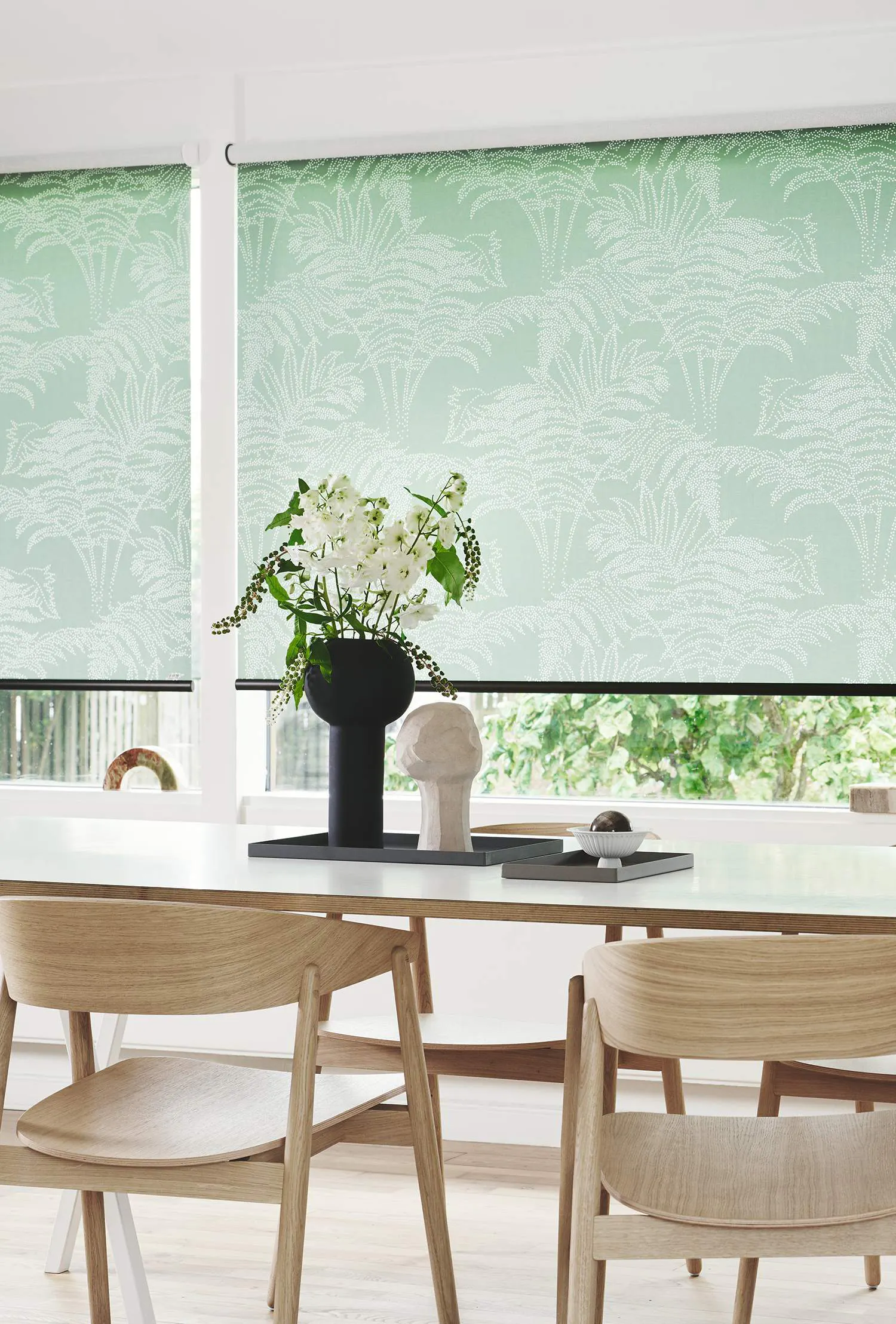 Ett modernt matrum med ljusa trämöbler och gröna rullgardiner med ett mönster av vita palmblad. På bordet står en svart vas med vita blommor och gröna blad, samt en dekorativ skulptur. I bakgrunden syns ett fönster som vetter mot en grönskande trädgård.