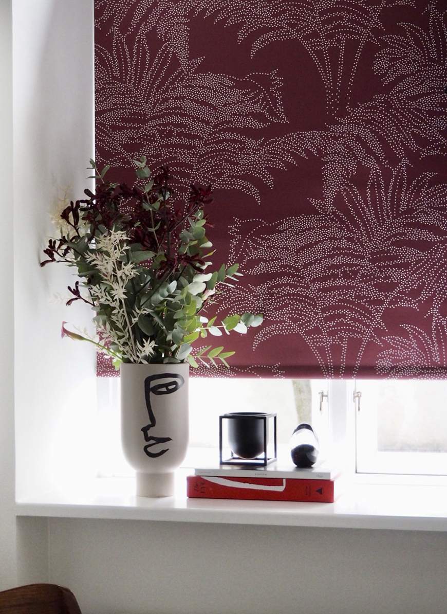 Ett fönsterbräde med dekorativa föremål: en vas med minimalistiskt ansiktsmotiv innehåller blommor och grönt, bredvid står en svart ljushållare och två staplade böcker – en röd och en vit. Fönstret har en vinröd rullgardin med ett prickigt palmbladsmönster, och naturligt ljus strömmar in genom fönstret.