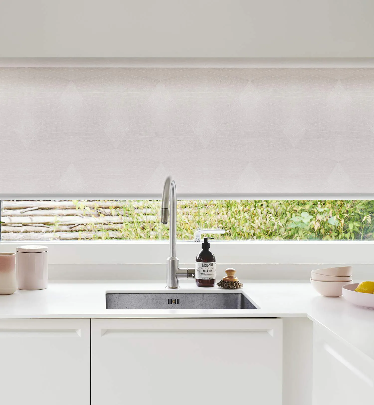 Ett modernt köksområde med en rostfri diskho och en hög, böjd kran. Till vänster om diskhon står två beige behållare, och till höger finns en flaska handtvål och en liten diskborste. Längre till höger står två skålar, varav en innehåller citroner. Fönstret bakom har en ljus rullgardin delvis neddragen, och utanför syns grönska samt en trästaket eller vägg.