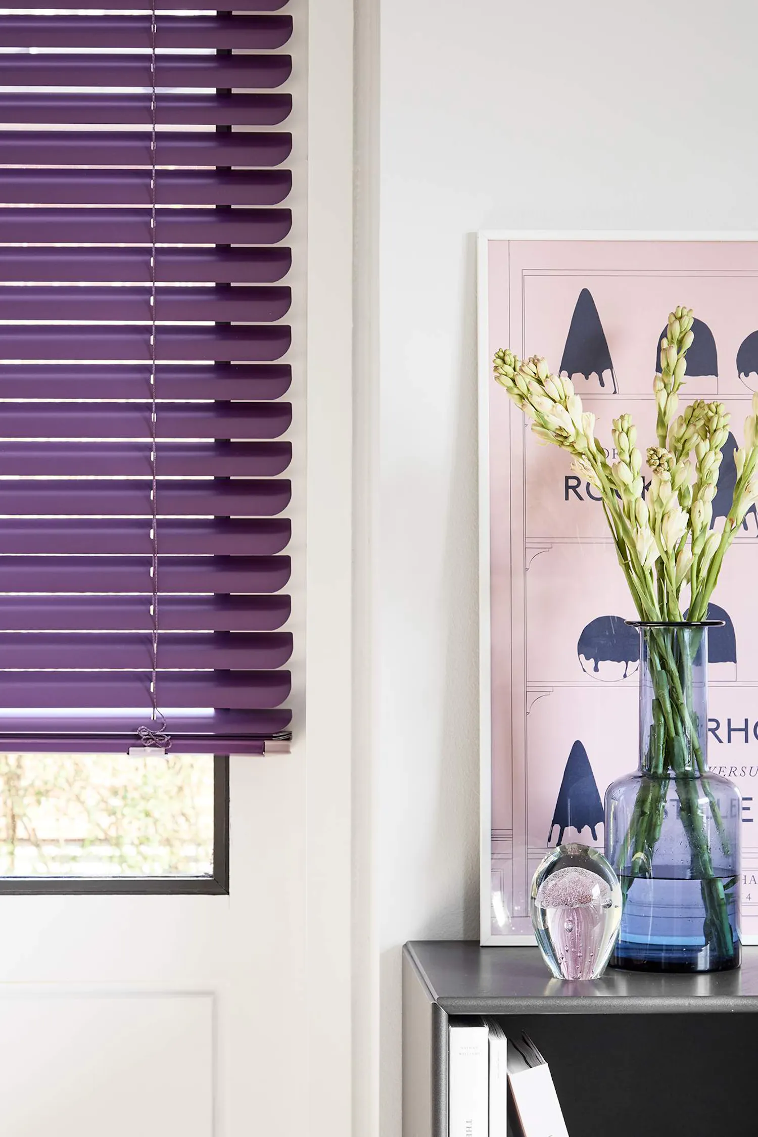 En bild som visar ett rum med lila persienner på ett fönster till vänster. Till höger finns en hylla med en vas fylld med vita blommor, en mindre glasprydnad och några böcker. Bakom vasen står en inramad affisch lutad mot väggen.