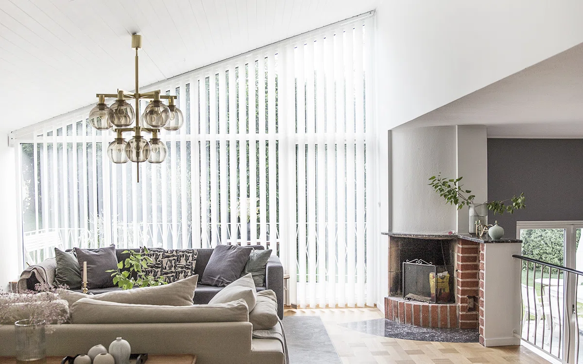 Ett modernt vardagsrum med högt i tak och stora vertikala fönster täckta av vita persienner. Rummet har en beige hörnsoffa med flera kuddar, ett soffbord med dekorativa föremål och en tegelspis till höger. Ovanför soffan hänger en elegant ljuskrona med flera glaskupor. Rummet är ljust tack vare naturligt ljus från fönstren, och några växter ger grönska till inredningen.