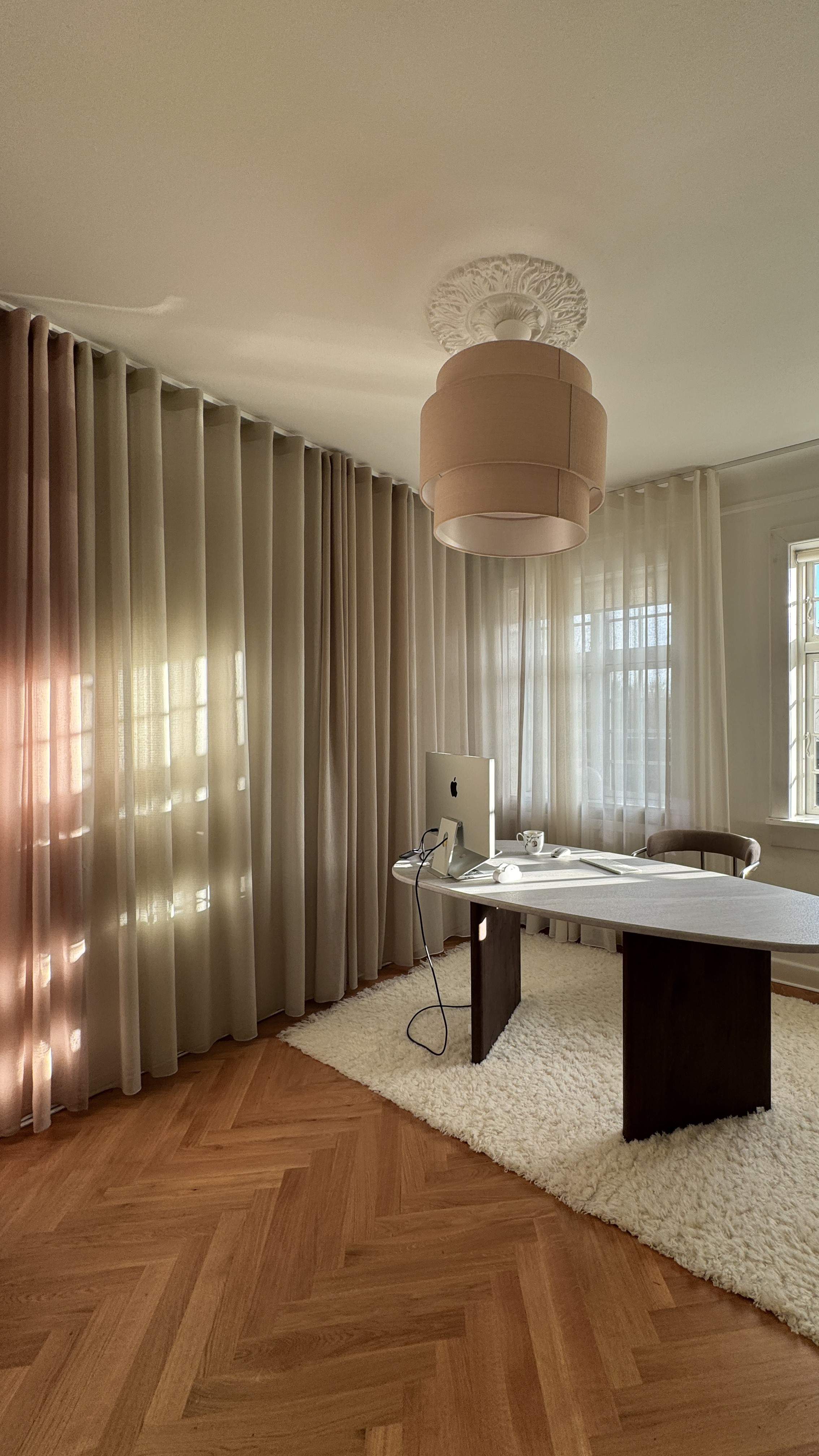 Moderne og minimalistisk kontor med store vinduer dekket av lyse gardiner som slipper inn naturlig lys. Et trebord med dataskjerm, kabler og tilbehør står på et gulv med fiskebensmønstret parkett. Under bordet ligger et hvitt, fluffy teppe. En unik taklampe henger over arbeidsområdet og gir rommet et stilrent og rolig uttrykk.
