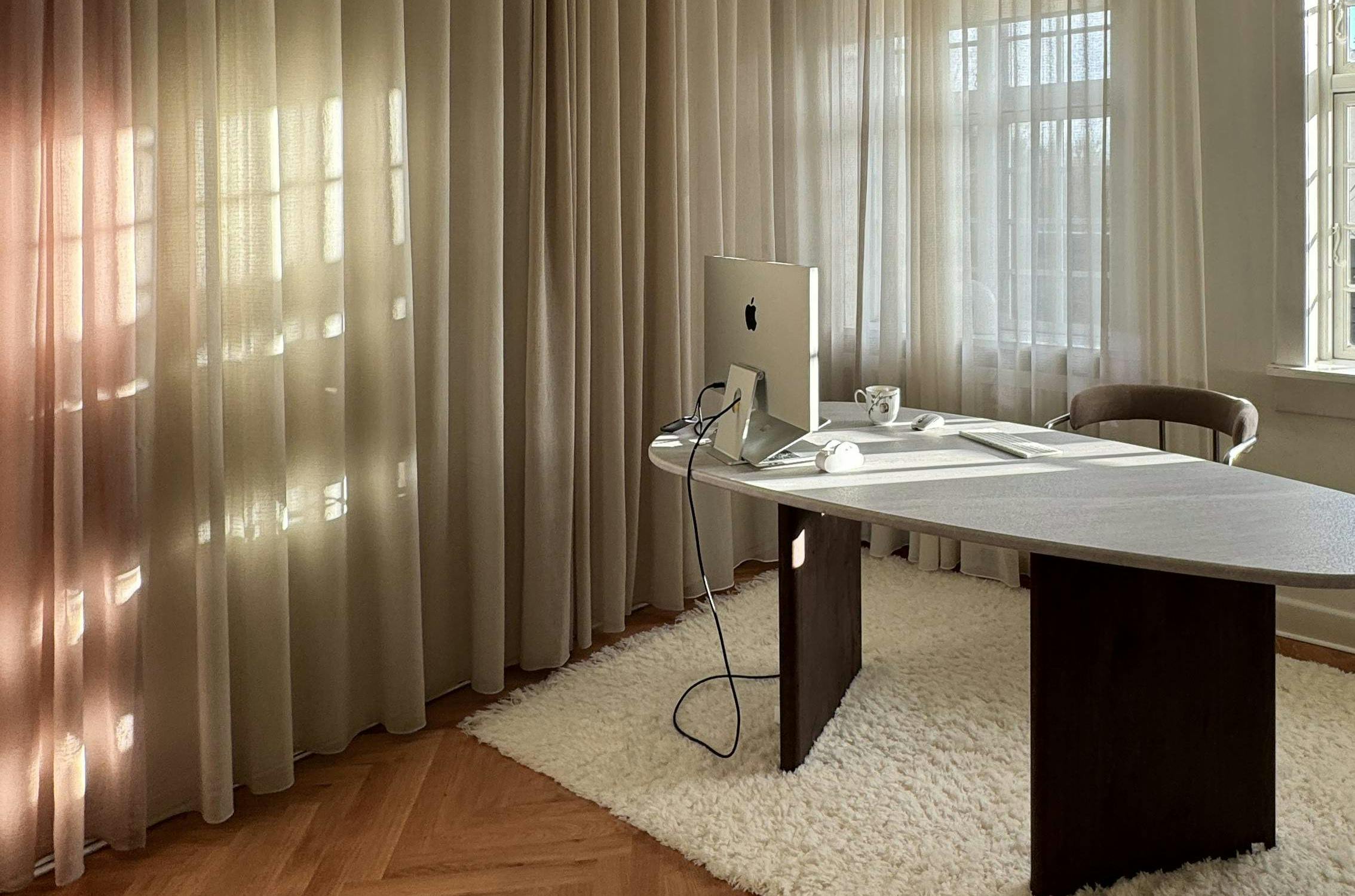 Moderne og minimalistisk kontor med store vinduer dekket av lyse gardiner som slipper inn naturlig lys. Et trebord med dataskjerm, kabler og tilbehør står på et gulv med fiskebensmønstret parkett. Under bordet ligger et hvitt, fluffy teppe. En unik taklampe henger over arbeidsområdet og gir rommet et stilrent og rolig uttrykk.
