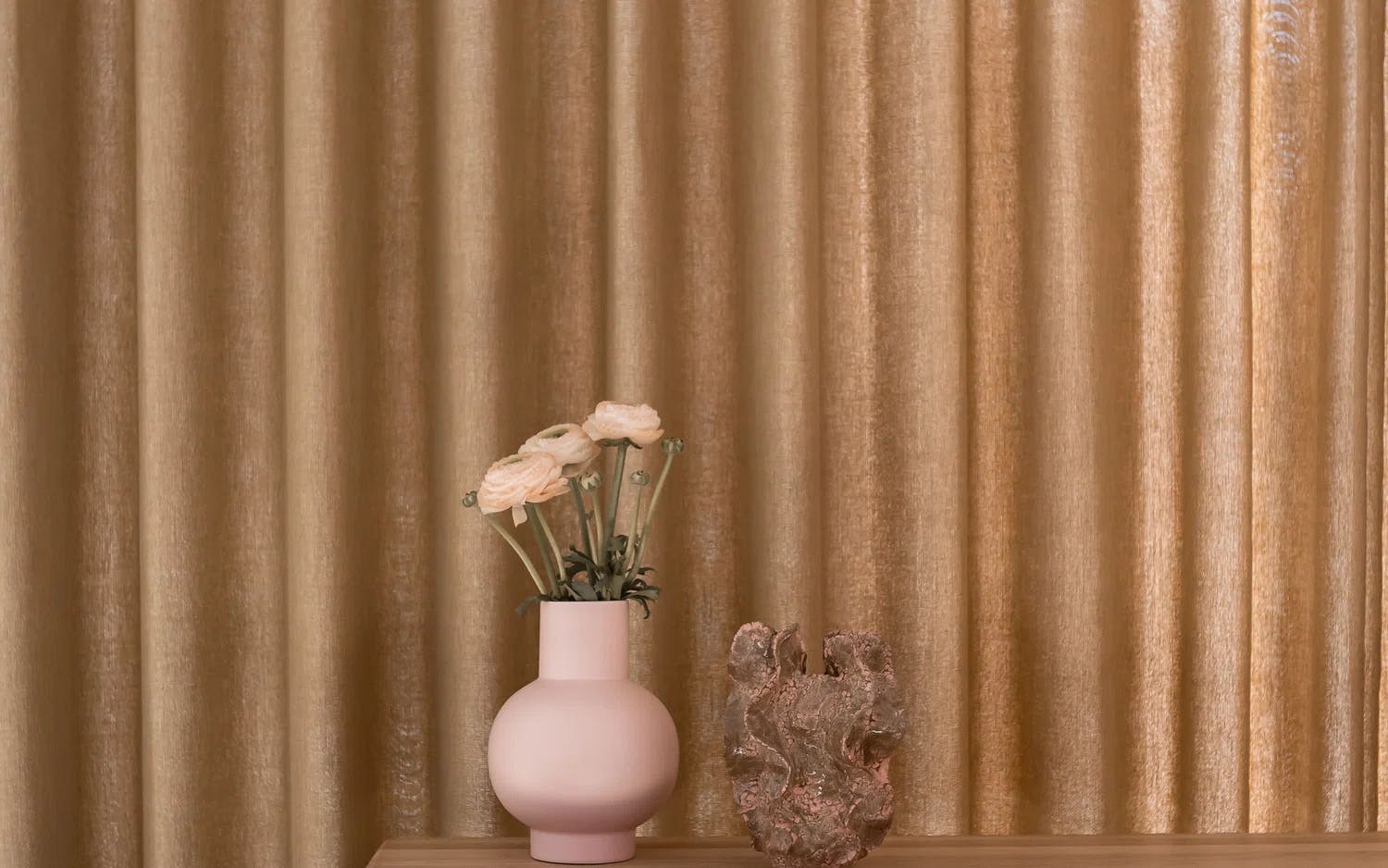 Rosa vase med hvite blomster og abstrakt skulptur foran gyldne gardiner – elegant og varm stemning.