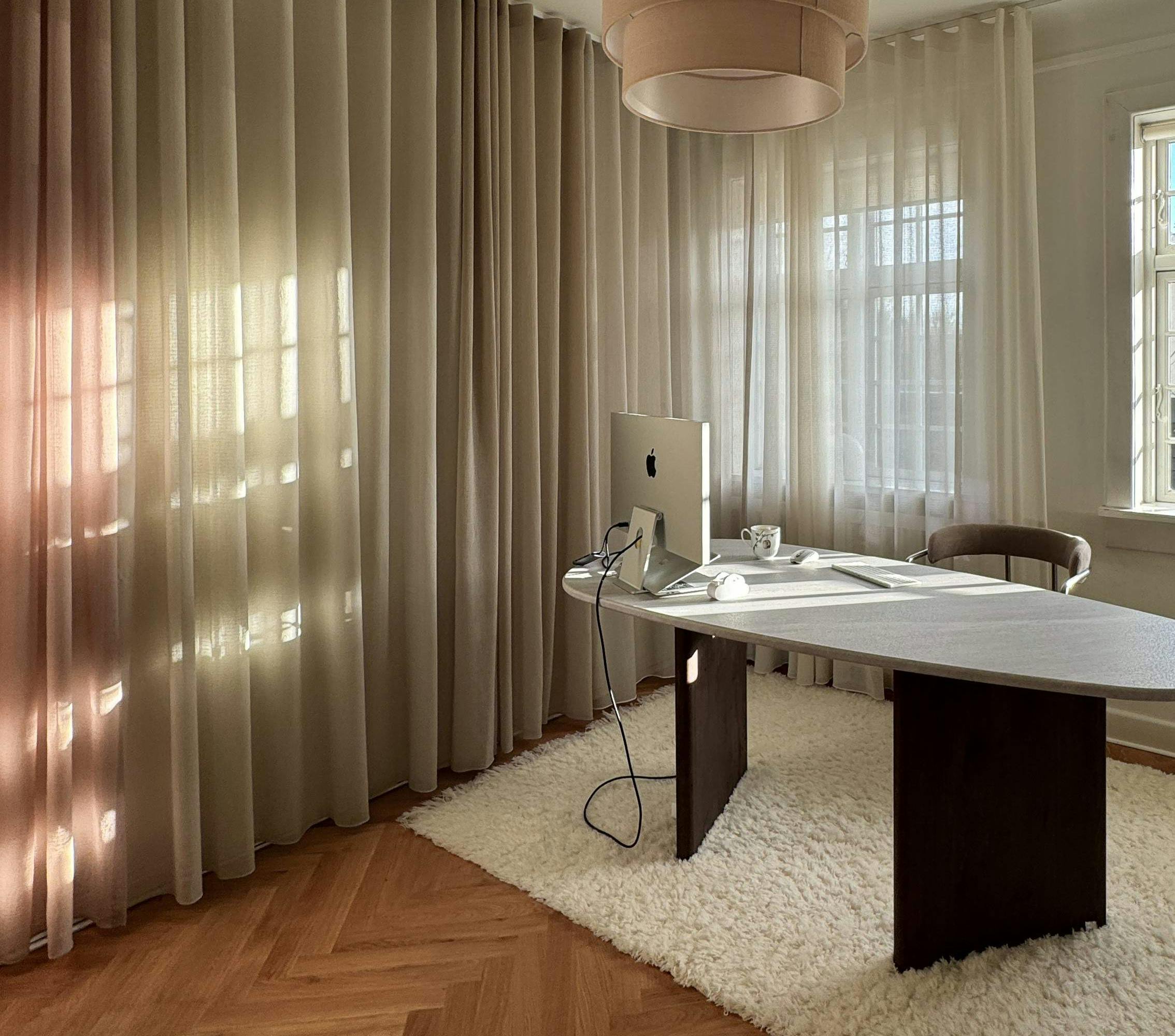 Modern minimalist arbetsplats med skrivbord, dator och mjuk belysning i ett ljust rum med gardiner och trägolv.