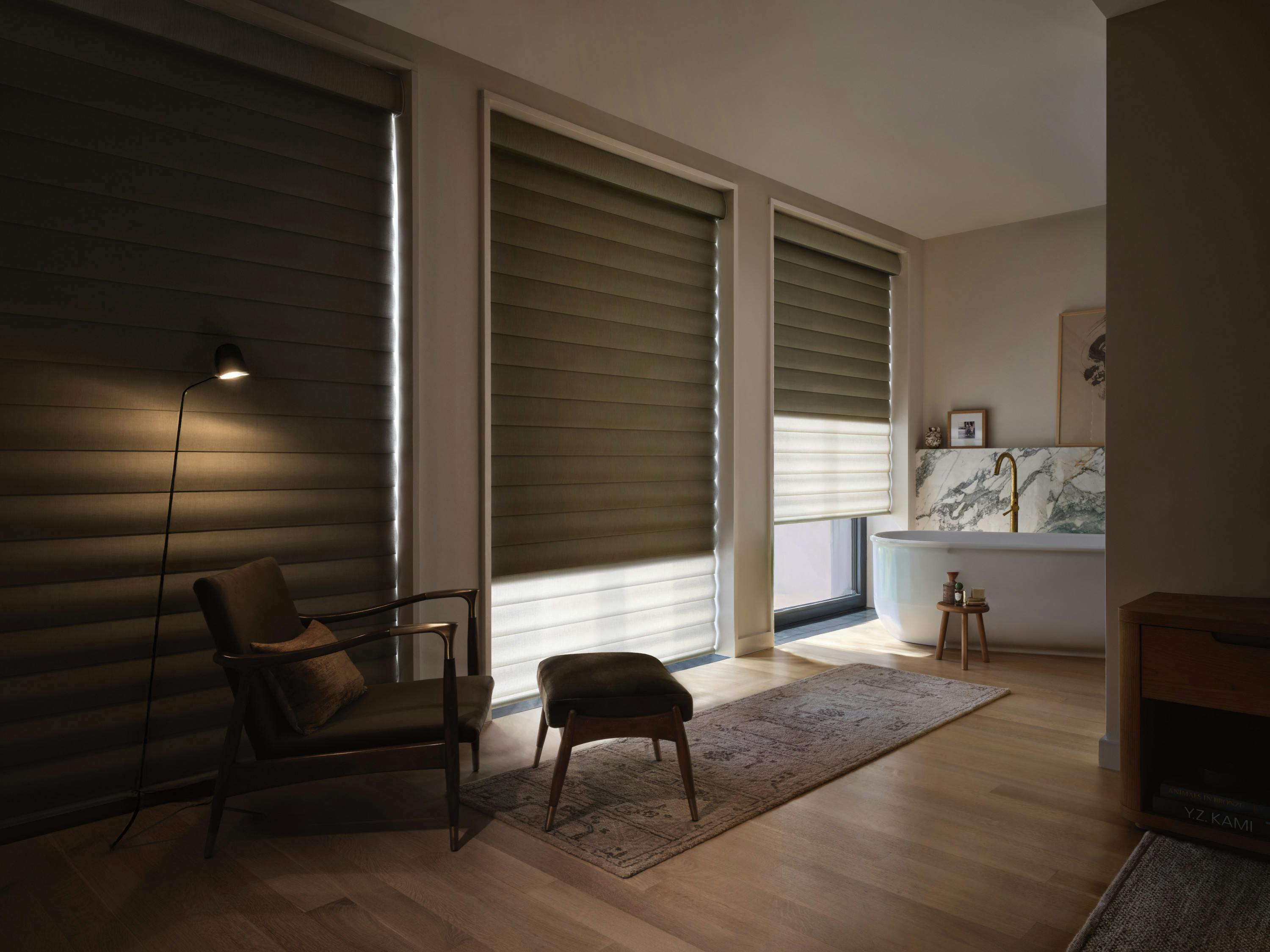 Cozy modern room with three large windows covered by dark layered shades, an armchair with a matching ottoman, a floor lamp, and a freestanding bathtub in the corner.