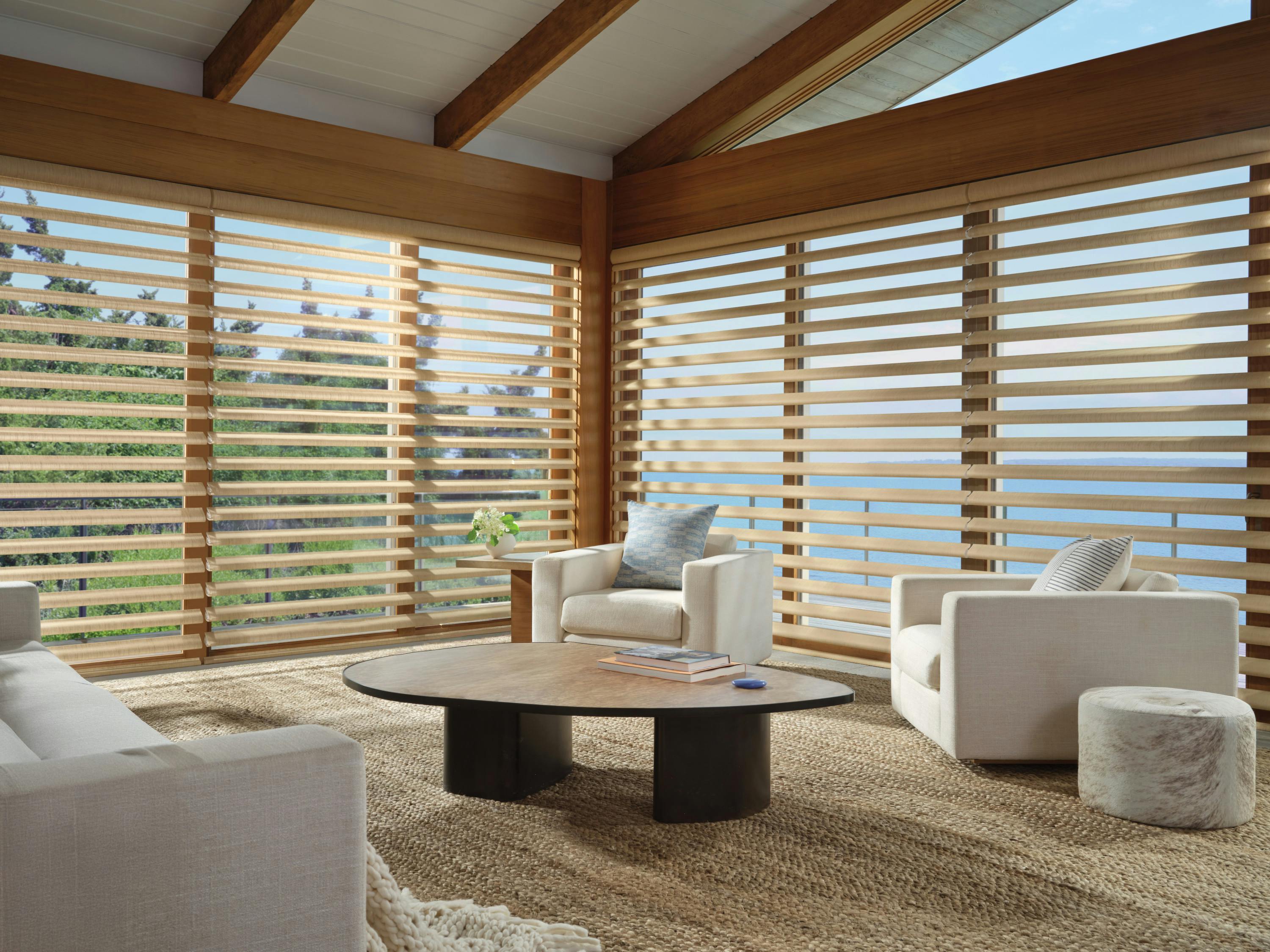 Bright coastal living room with floor-to-ceiling wooden-framed windows featuring horizontal layered shades, two white armchairs, a white sofa, a round wooden coffee table, and a textured rug.