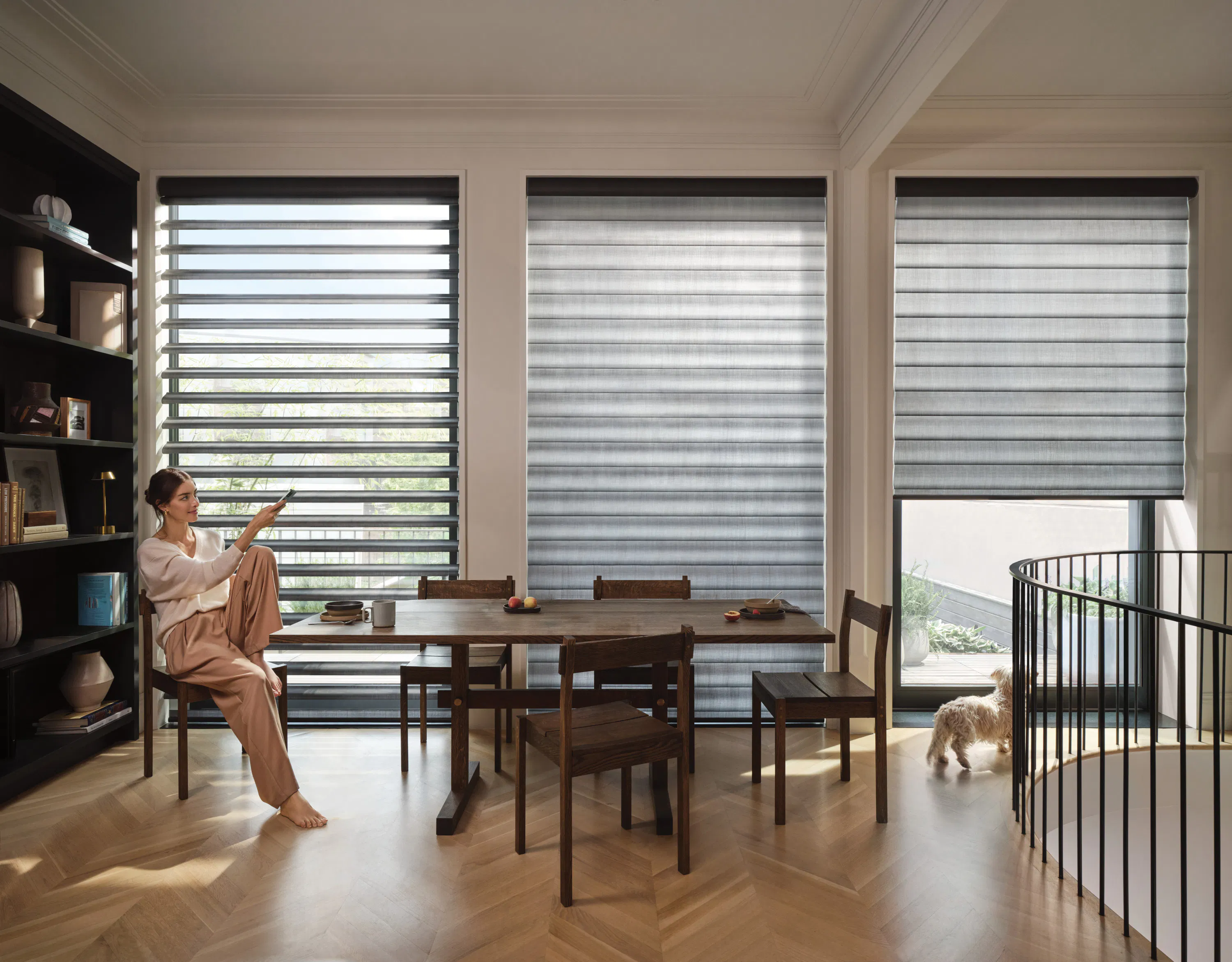 Dining area with three large windows featuring dark layered shades, a wooden table with chairs, open shelving on the left, and a small dog standing near a staircase railing.