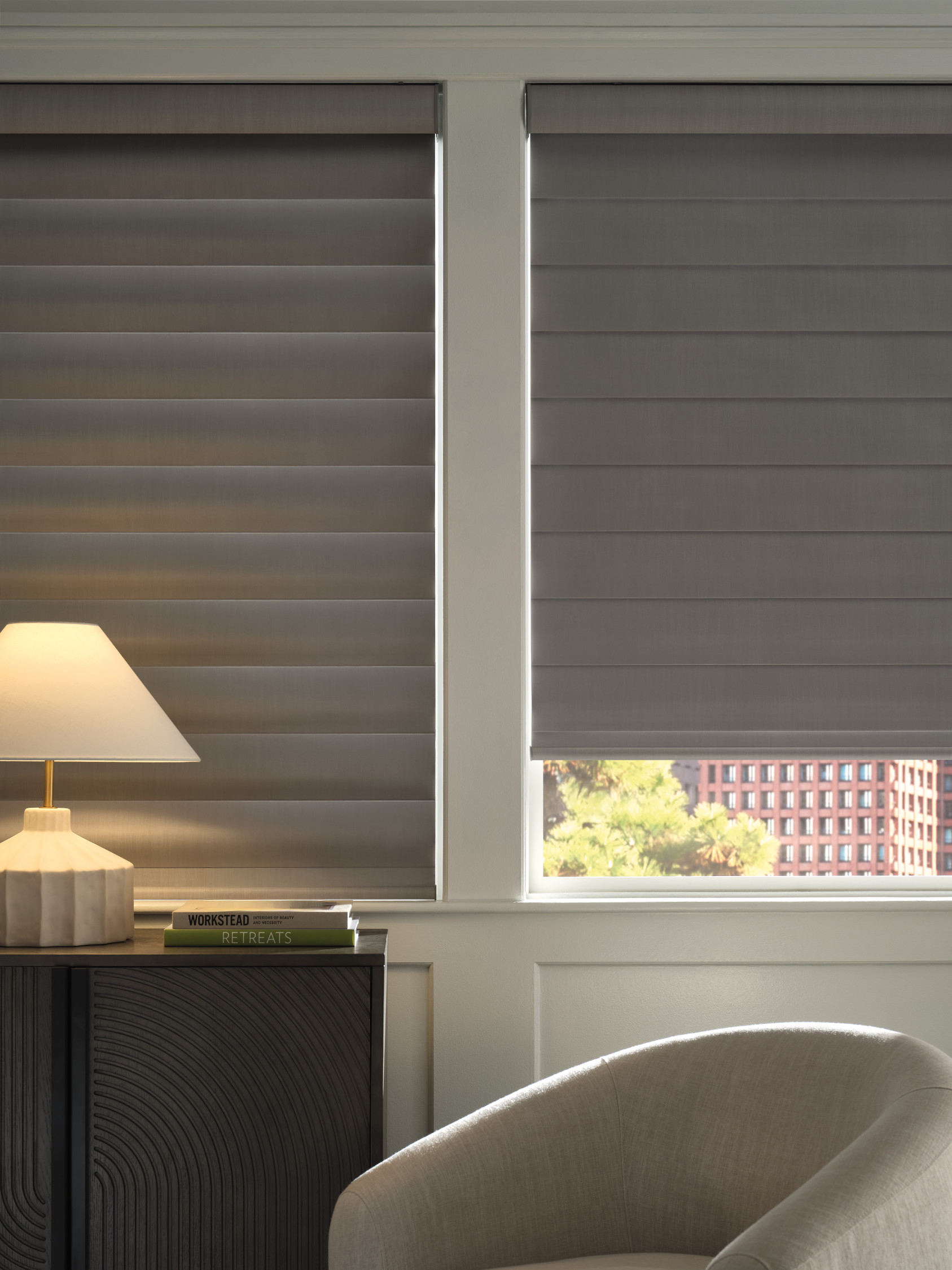 Close-up of two windows with grey layered shades, a lamp on a cabinet with books, and part of a cushioned chair in the foreground.