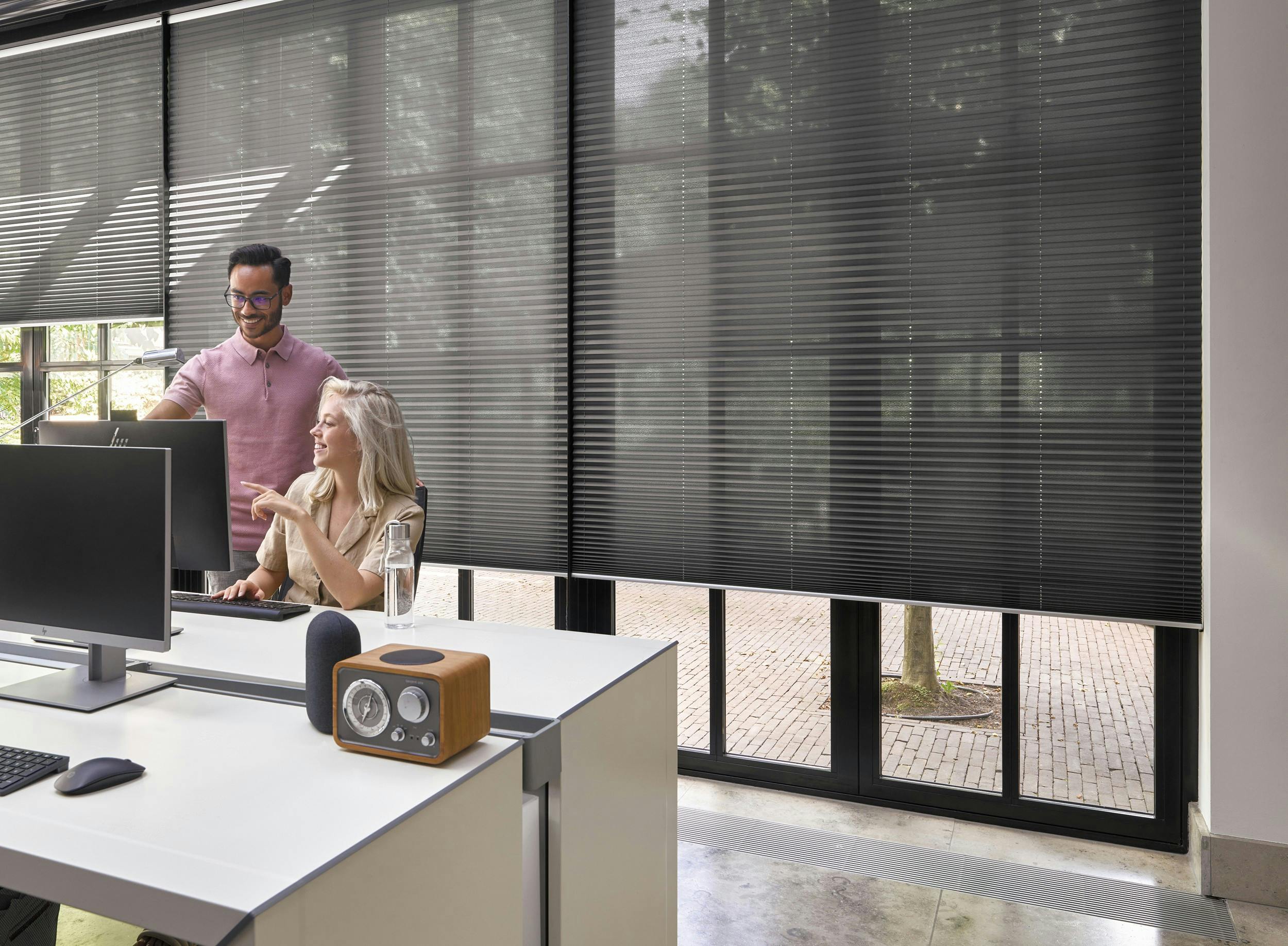 A man and woman in an office with black plisse shades on large windows