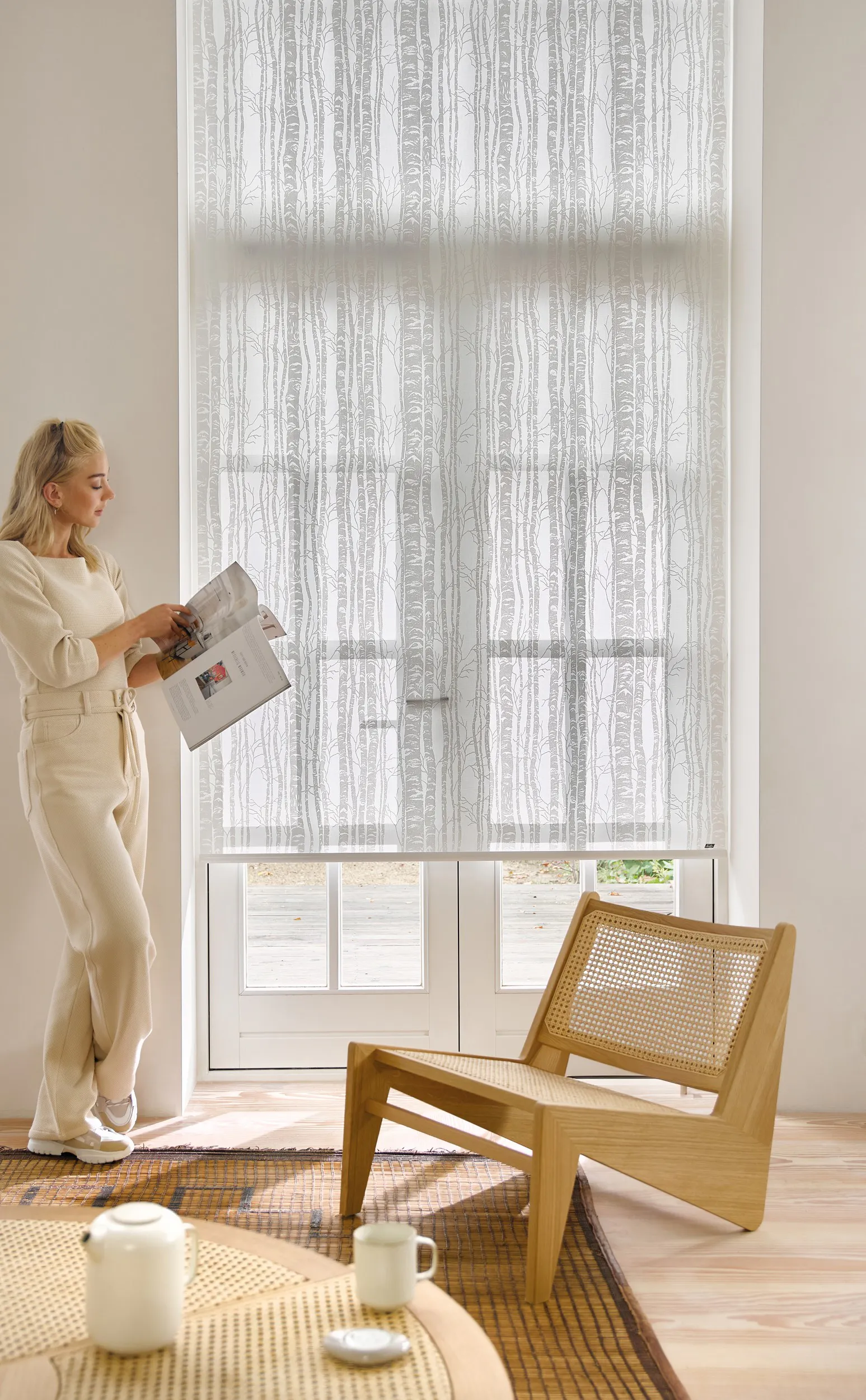 A woman reading magazine in a light living room with white roller blinds on patio doors
