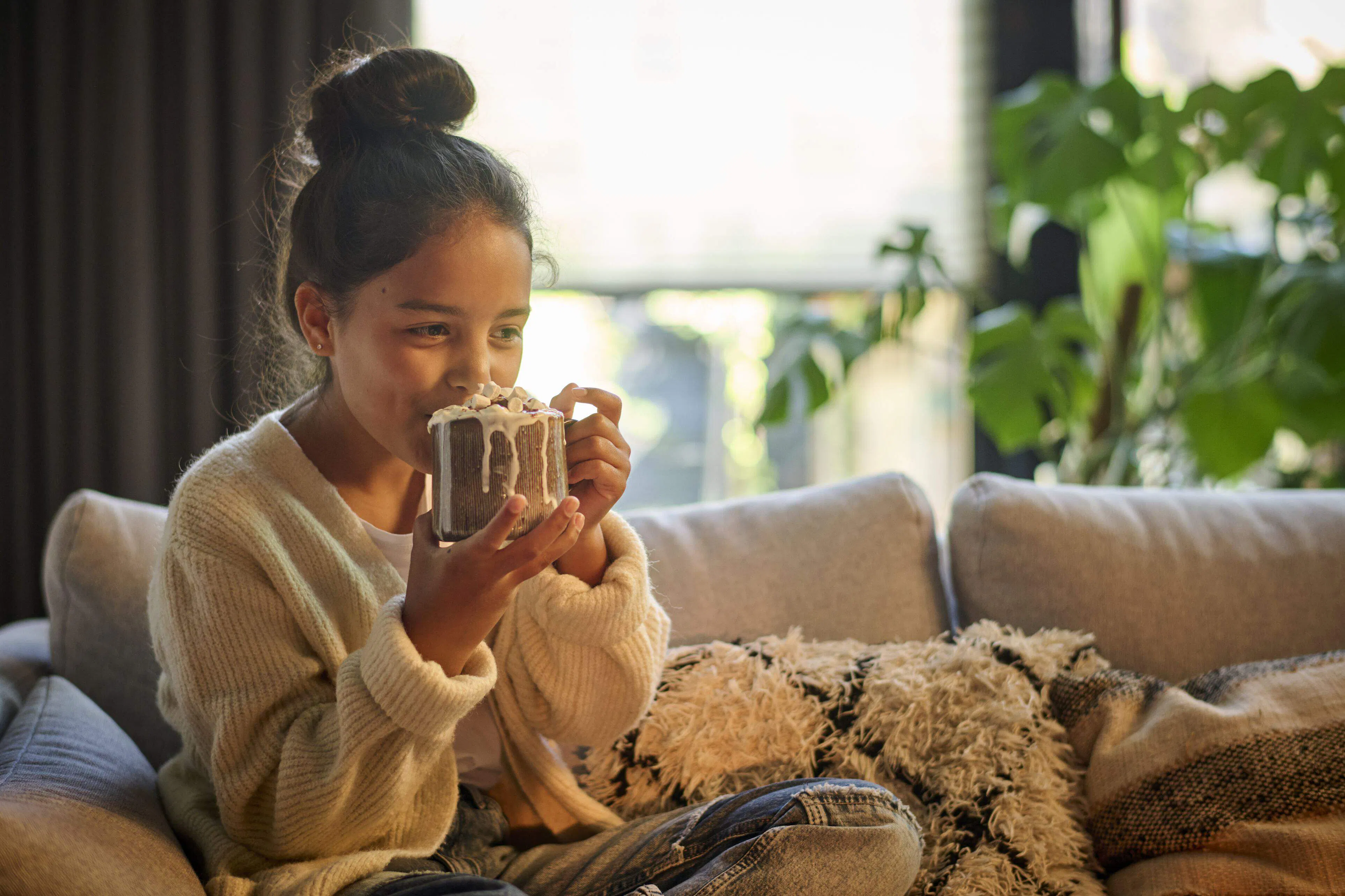 A girl drinking from a warm drink in a cozy living room with white duette blinds layered with brown curtains