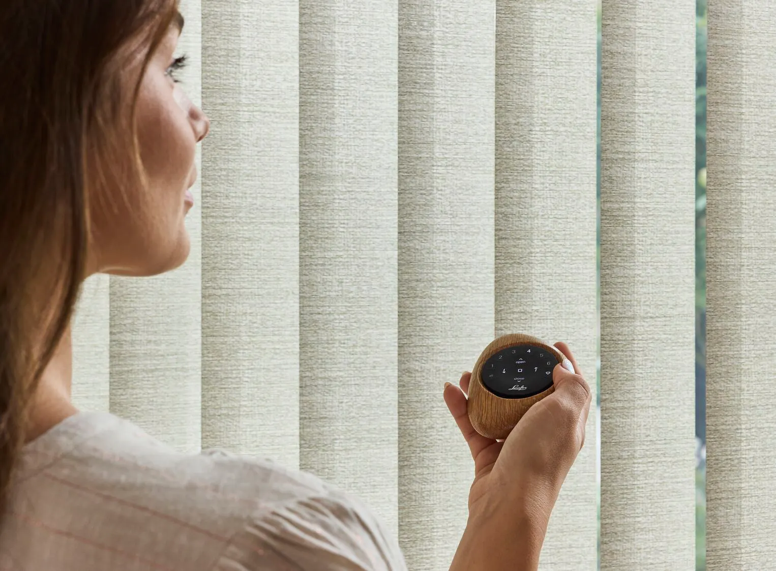 Gros plan d’une femme tenant une télécommande Pebble®, avec des stores verticaux beiges en arrière‑plan