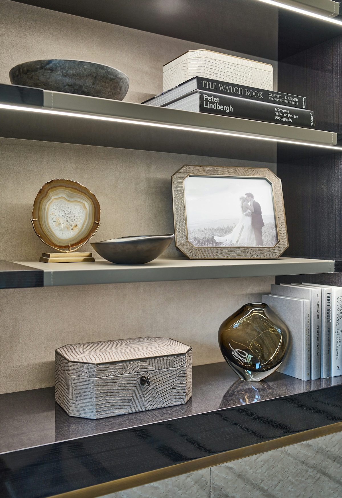 Modern open shelving with accessories including an agate slice, a bronze chamfered photo frame featuring a photo of a bride and groom. a matching jewellery box and a smoky glass vase holding up a set of books.