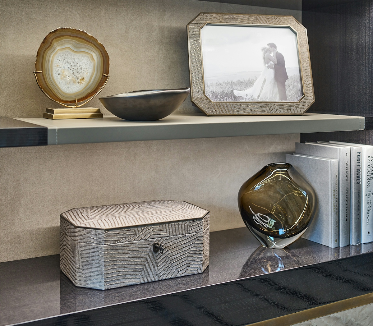 Modern open shelving with accessories including an agate slice, a bronze chamfered photo frame featuring a photo of a bride and groom. a matching jewellery box and a smoky glass vase holding up a set of books.