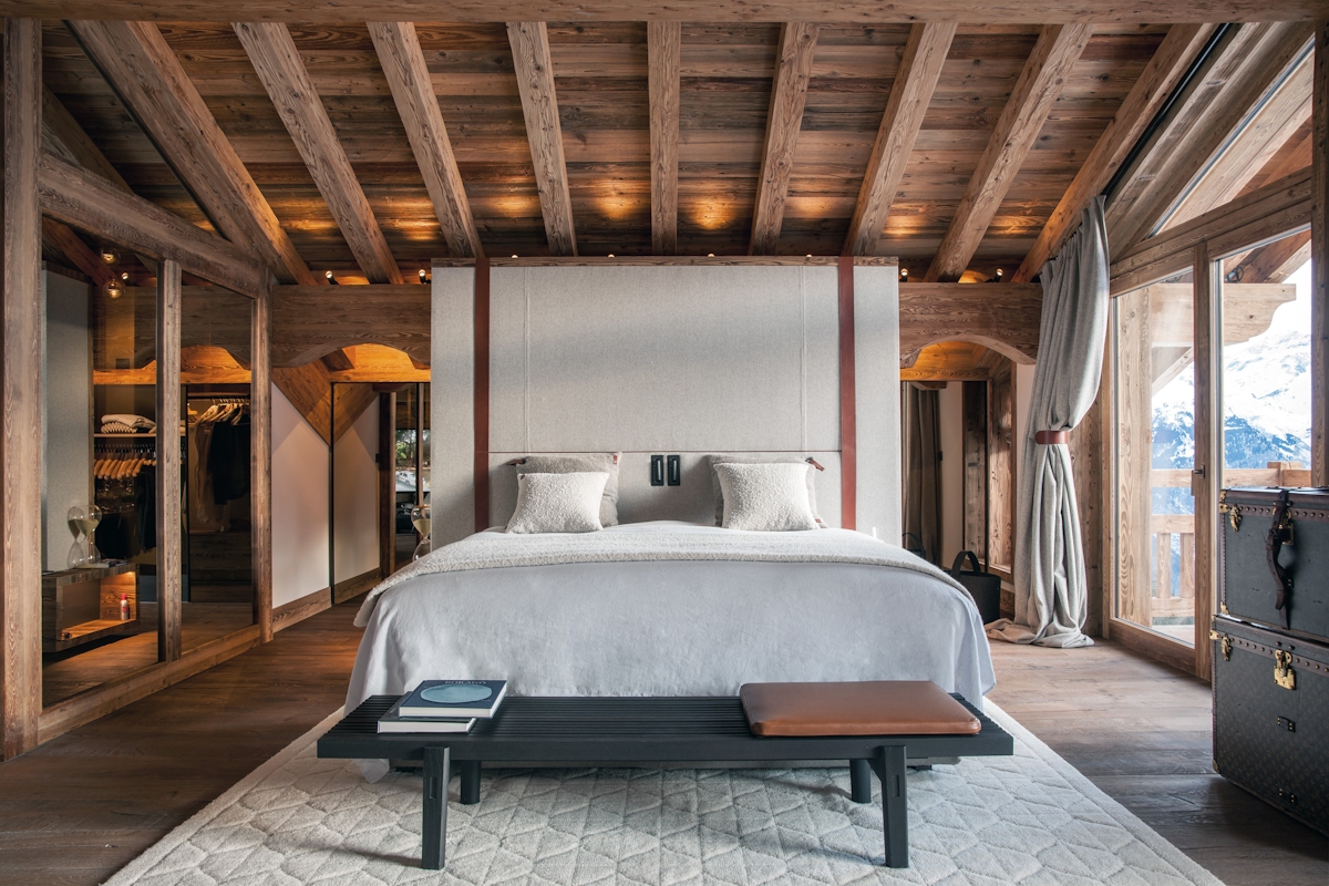 Atelier Giffon winter chalet bedroom with wooden beams and large white bed, black wooden bench, suitcase style storage.