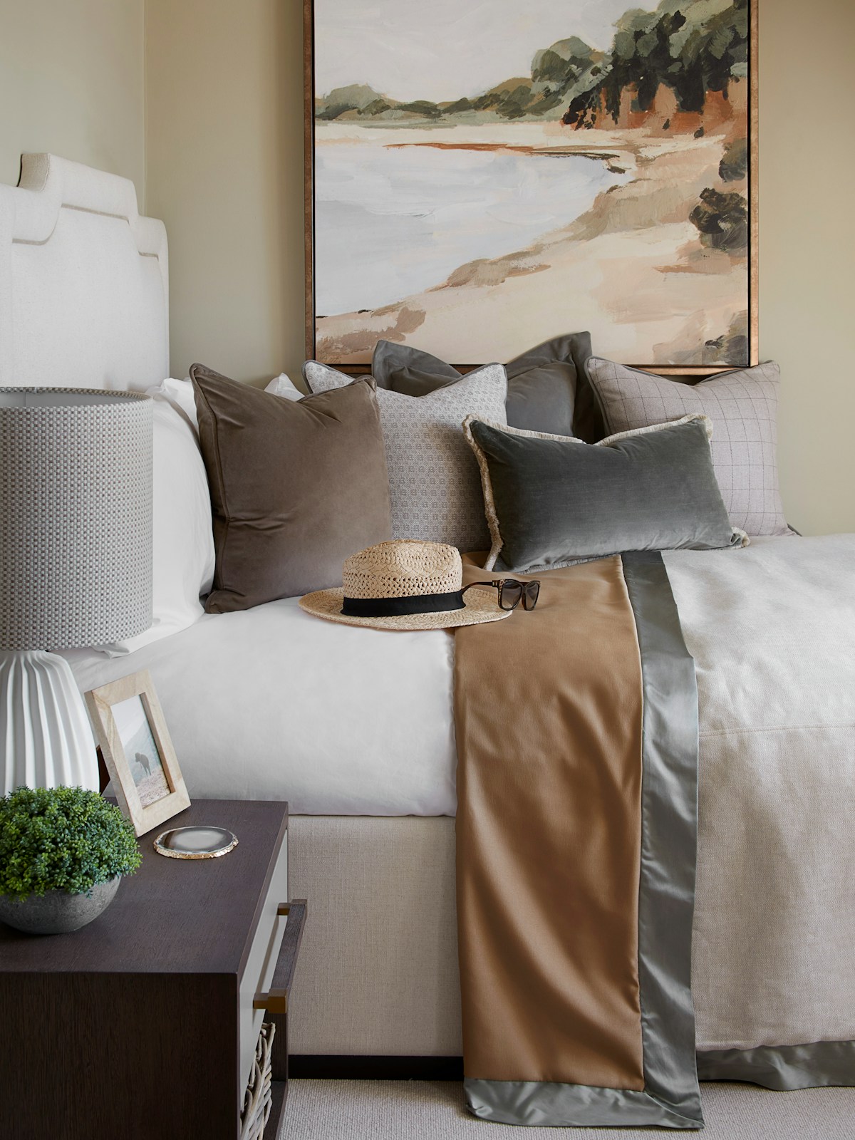 Inviting bedroom with Devereaux bed, "Bluff Bay" painting and various neutral cushions by LuxDeco, a small cream Teardrop porcelain table lamp by Jonathan Adler and 