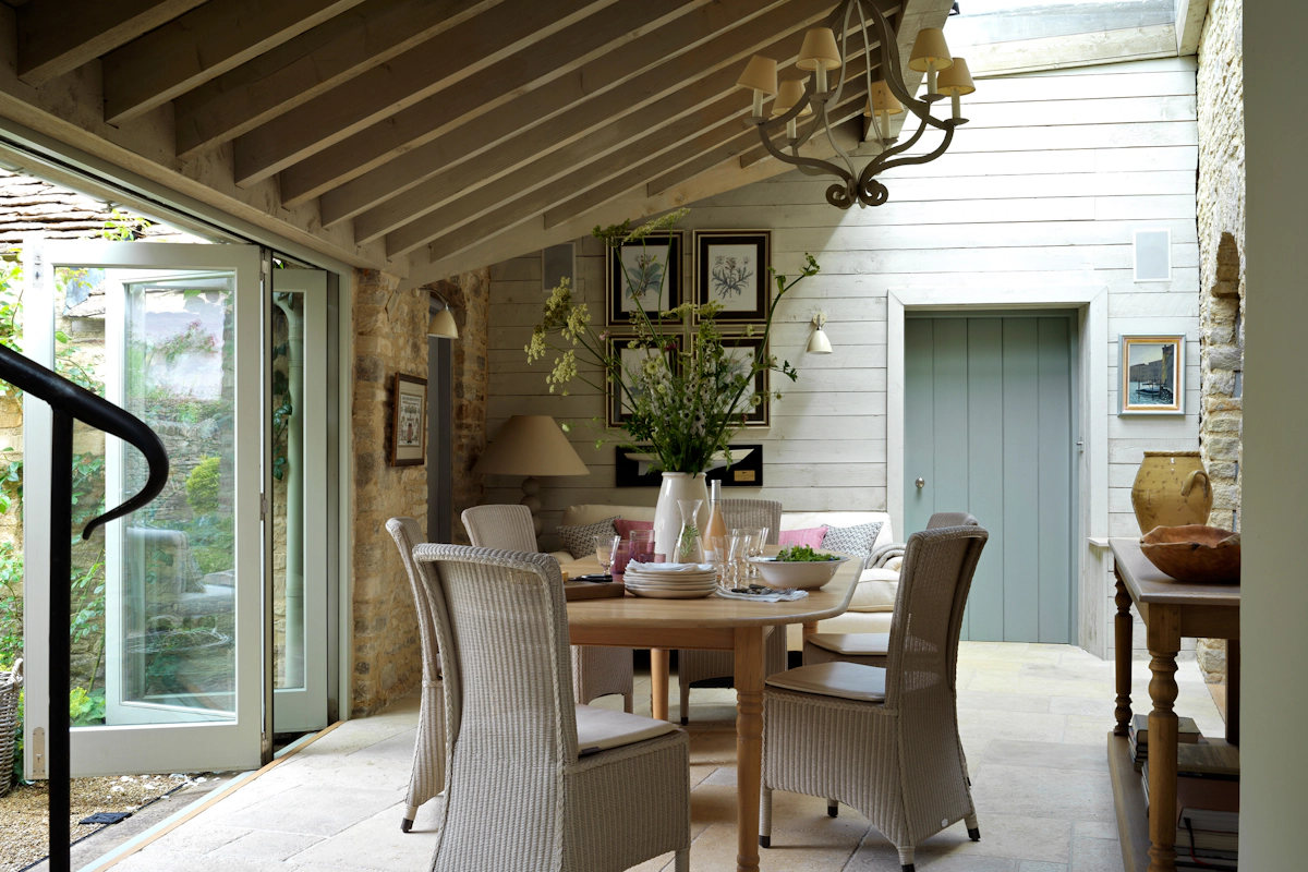 Emma Sims-Hilditch country style dining area with rattan dining chairs and wooden dining table. Stone floor and wooden beams feature with relaxed chandelier and wooden console table
