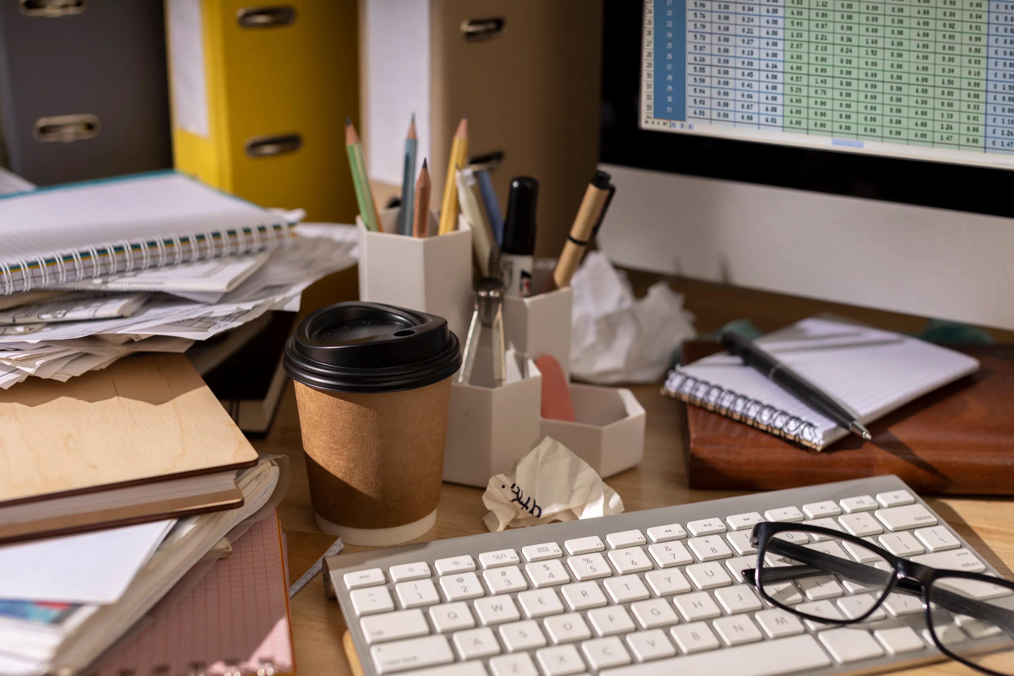 Cluttered desk stacked with paperwork and notebooks in front of a monitor showing a dense spreadsheet