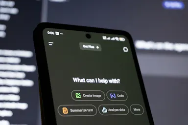 Smartphone held up to a code-filled monitor, showing an AI assistant asking what it can help with